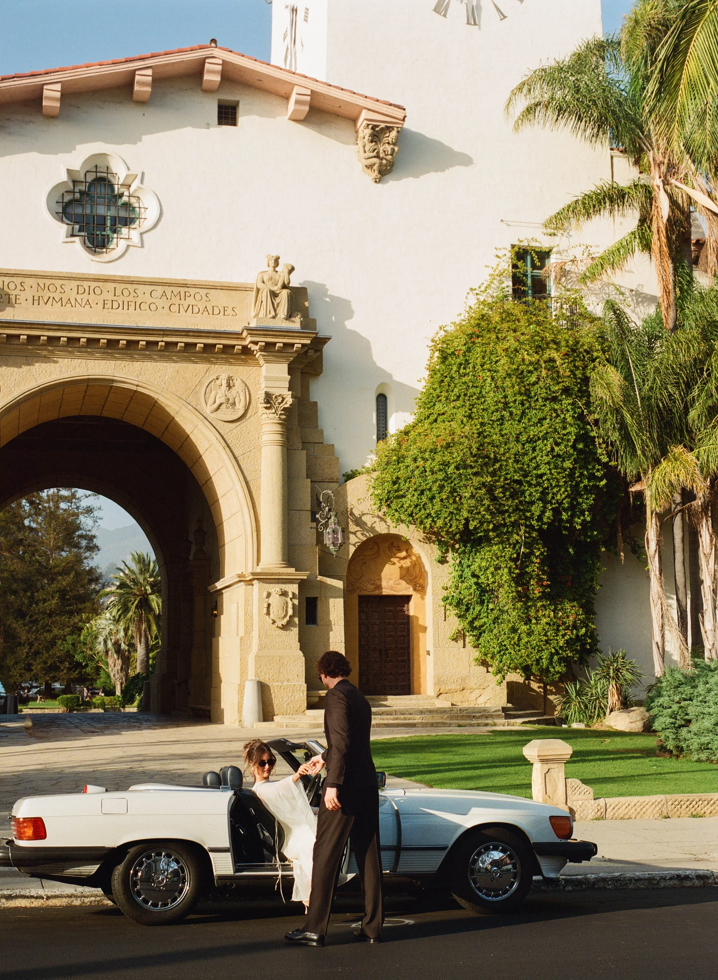 A woman in a white dress in a convertible car is being handed an item by a man in a black suit. They are in front of a historic building with an arched entrance, columns, and sculptures. There are palm trees and greenery around.