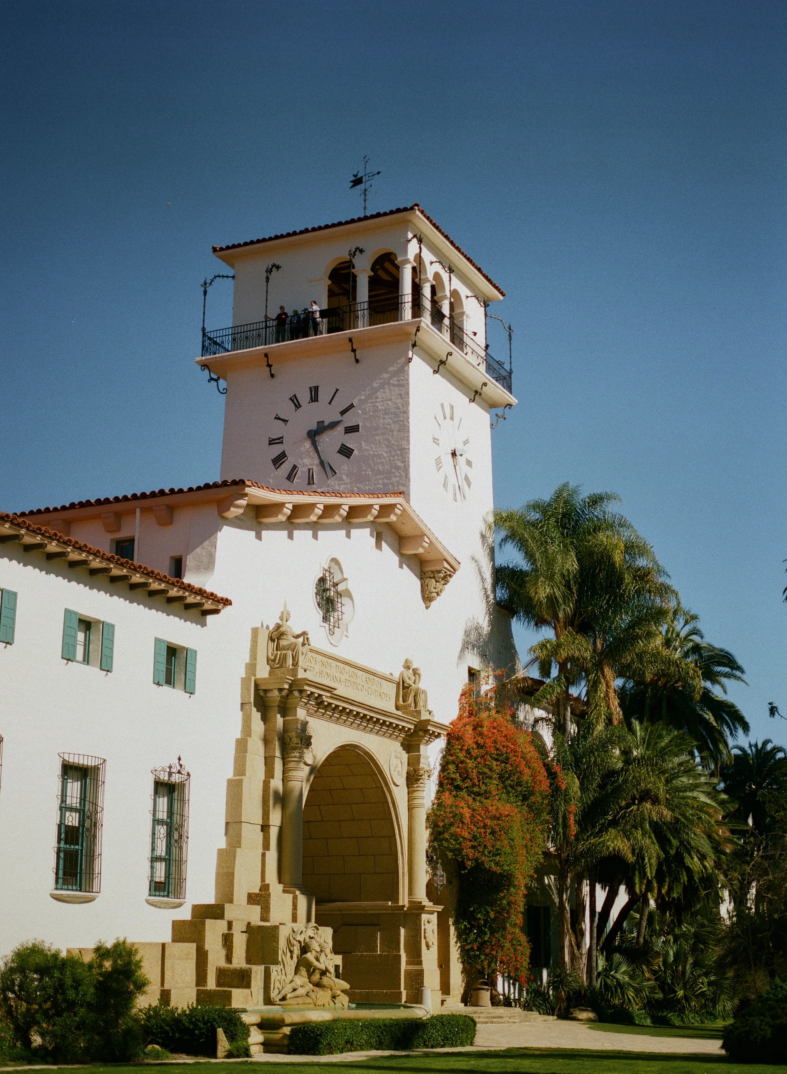 A clock tower with a white facade and Roman numeral clock faces, a decorative arched entrance, statuary, and lush landscaping including palm trees and flowering bushes.