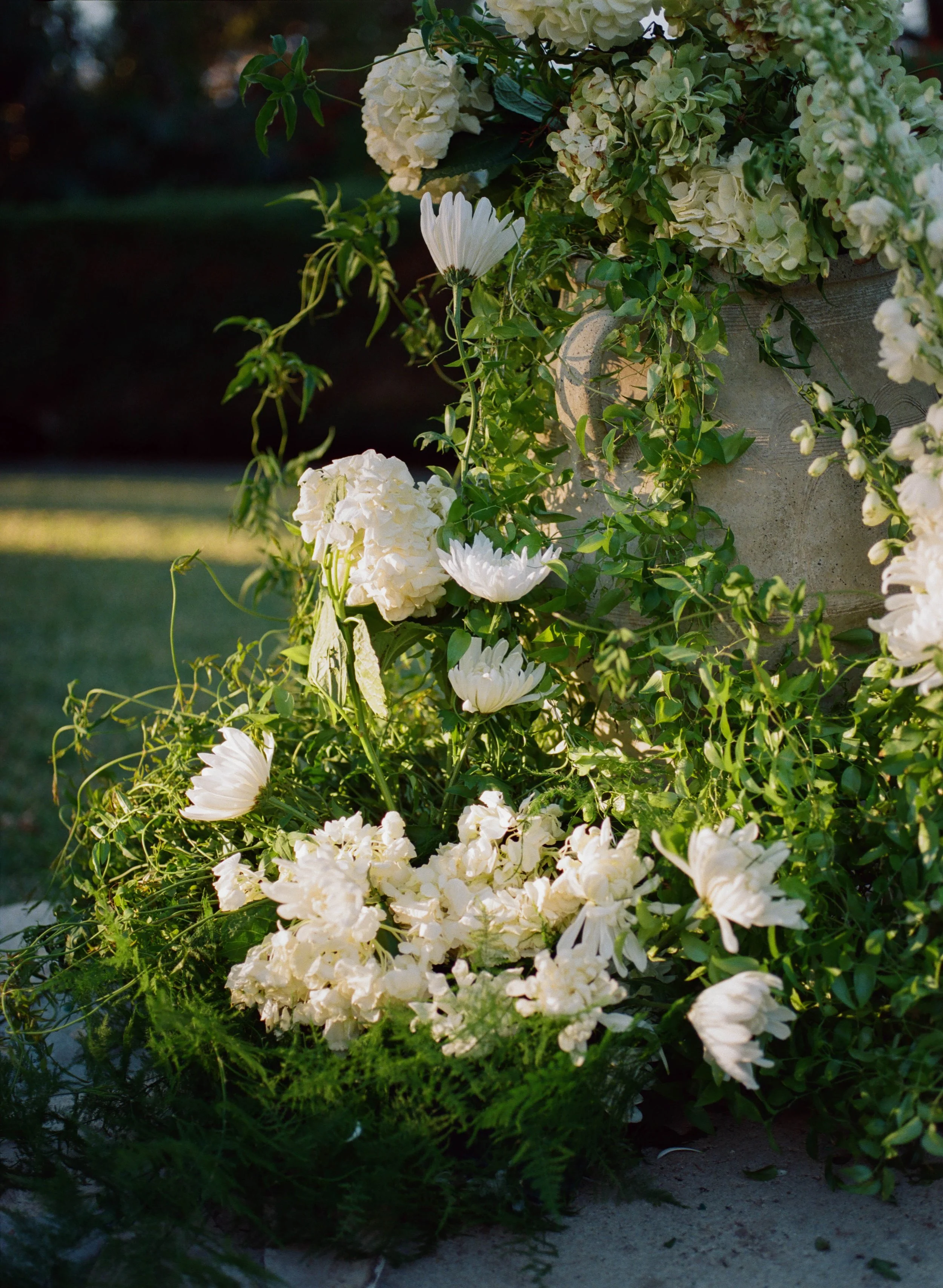 A floral arrangement with white hydrangeas, daisy-like flowers, and greenery in a concrete pot on a garden surface.