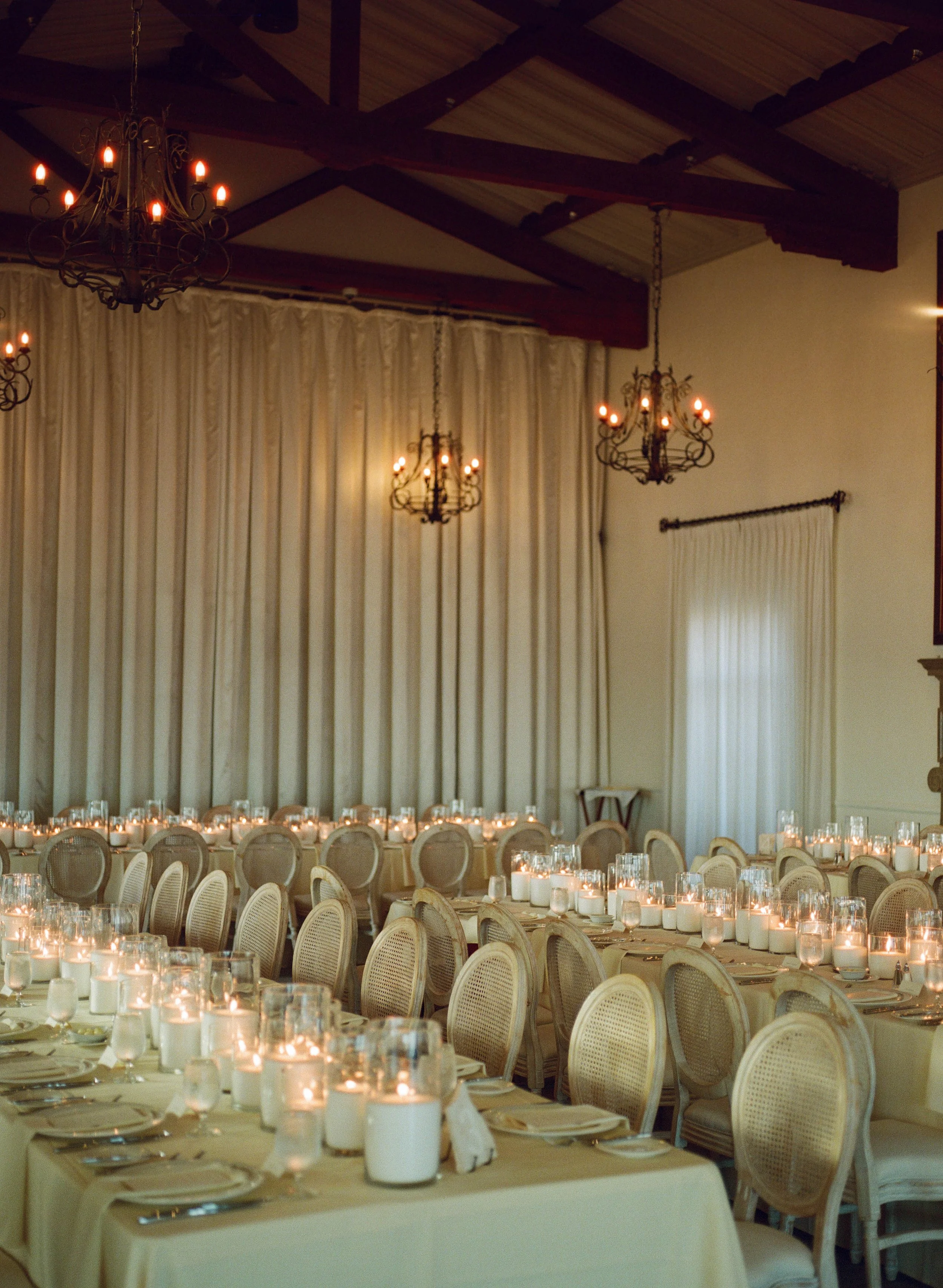 Elegant banquet hall with round tables, each decorated with multiple candles in glass holders, beige chairs, and cream-colored curtains in the background, illuminated by chandelier lights.
