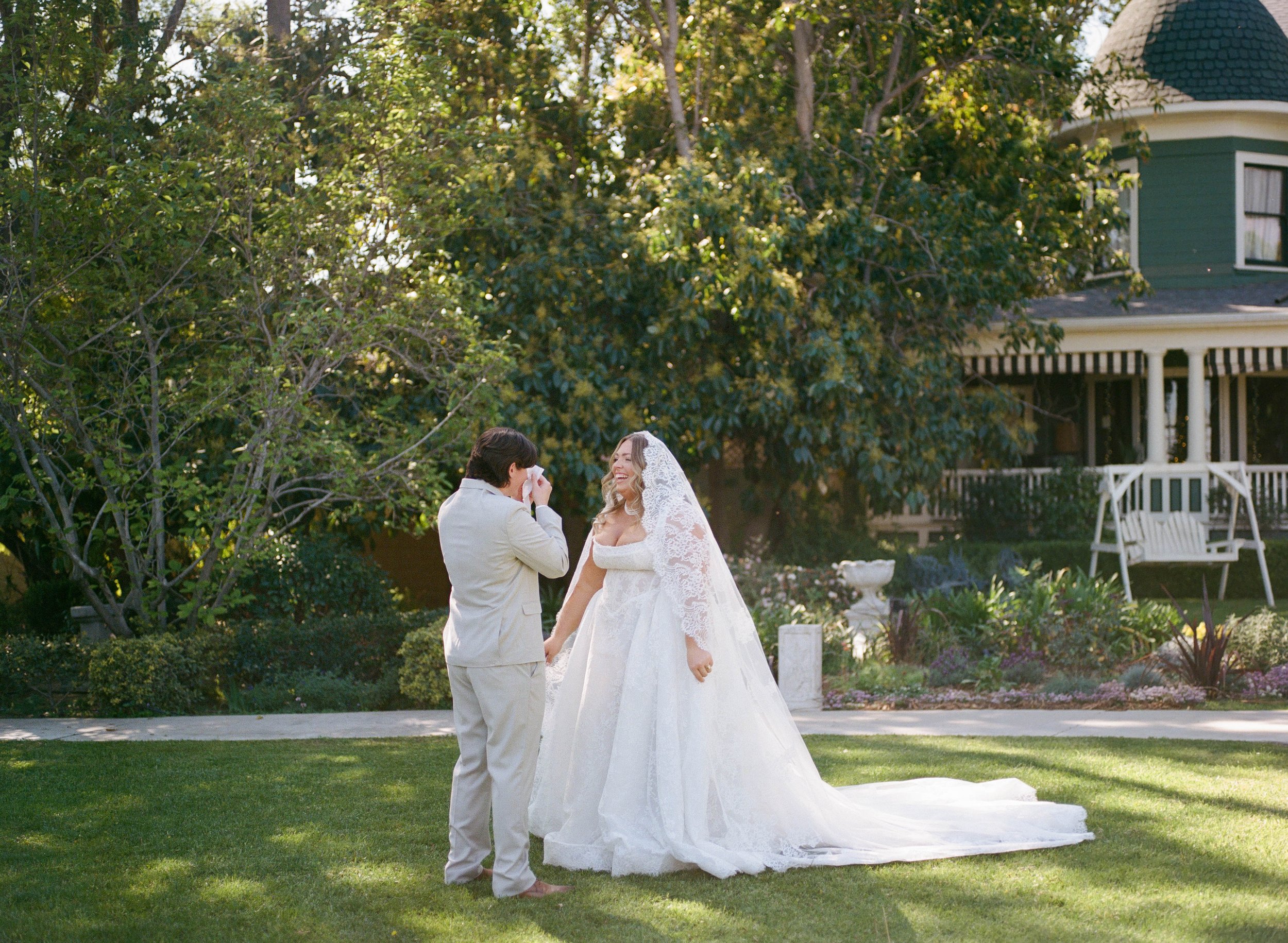 A wedding ceremony taking place outdoors in front of a green, leafy tree and a house with a porch. The bride, wearing a white lace wedding dress and veil, is smiling and holding hands with the groom, who is dressed in a light-colored suit.