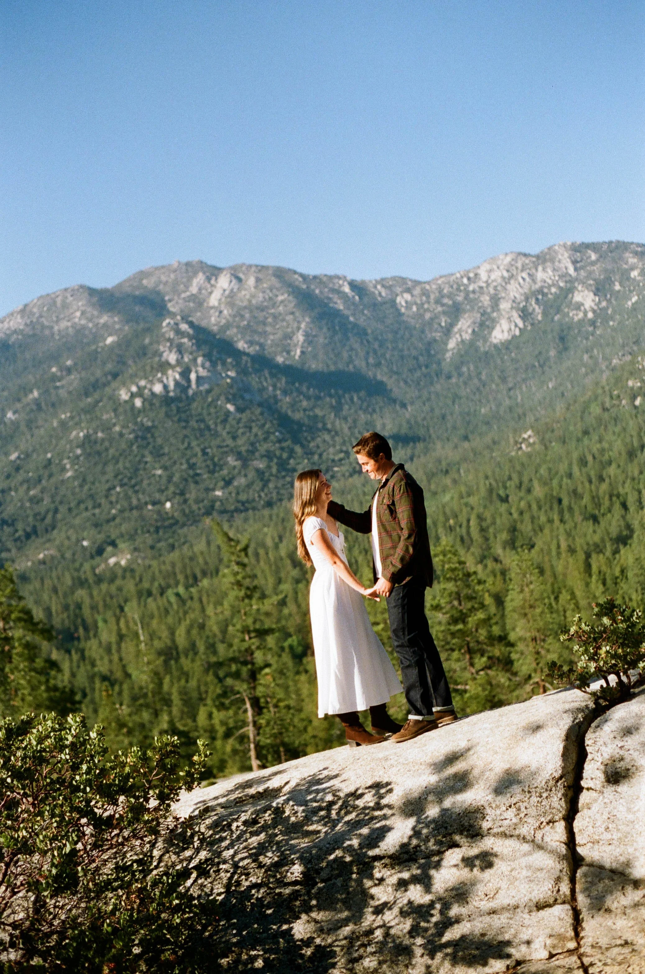 A couple stands on a rocky ledge holding hands, with a mountain covered in trees and a clear blue sky in the background.