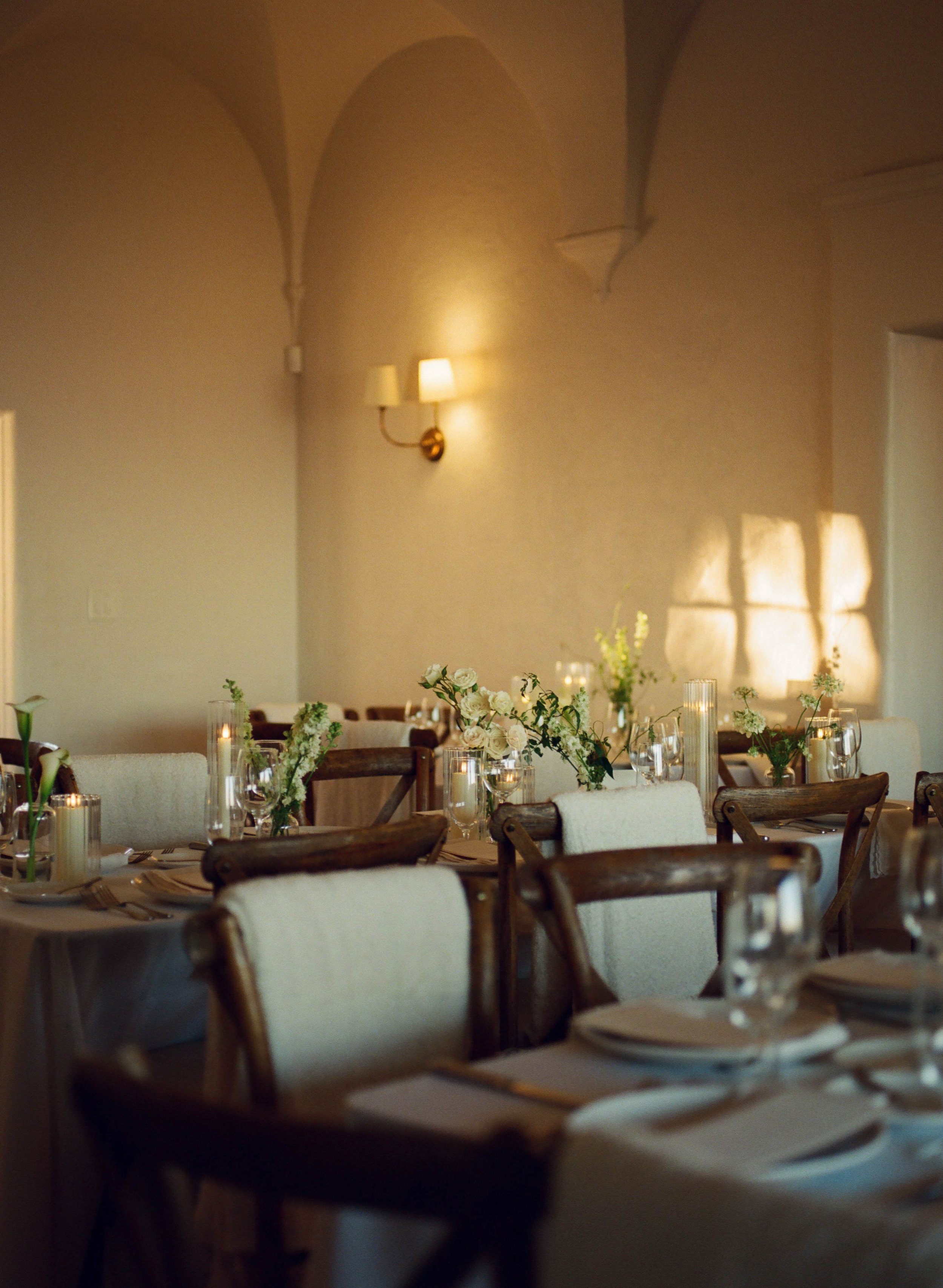A restaurant dining room decorated with white flowers, candles, and wine glasses on tables, with soft lighting and high ceilings with arches.