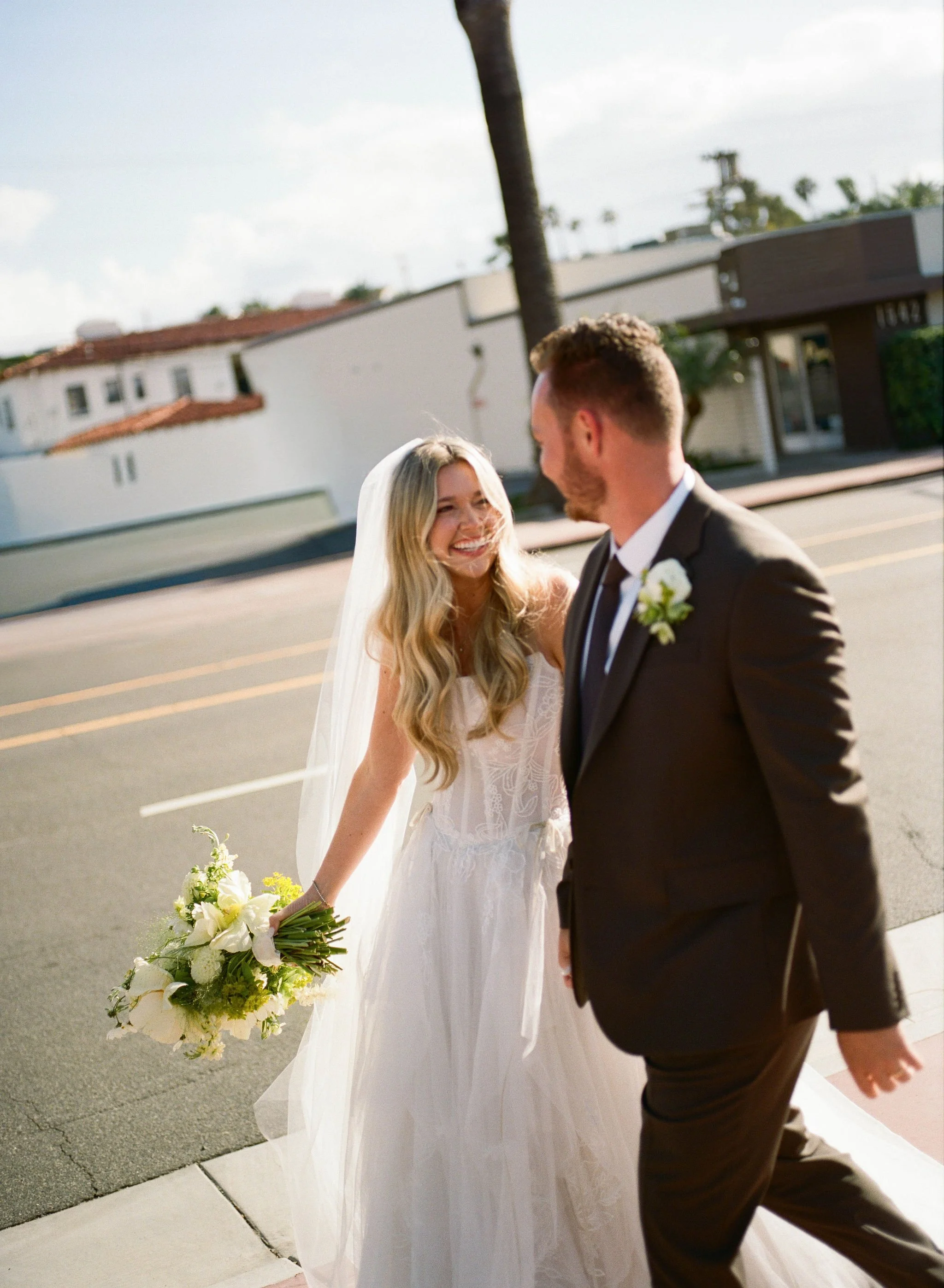 A bride holding a bouquet of white flowers smiling at a groom dressed in a black suit outside on a sunny day.