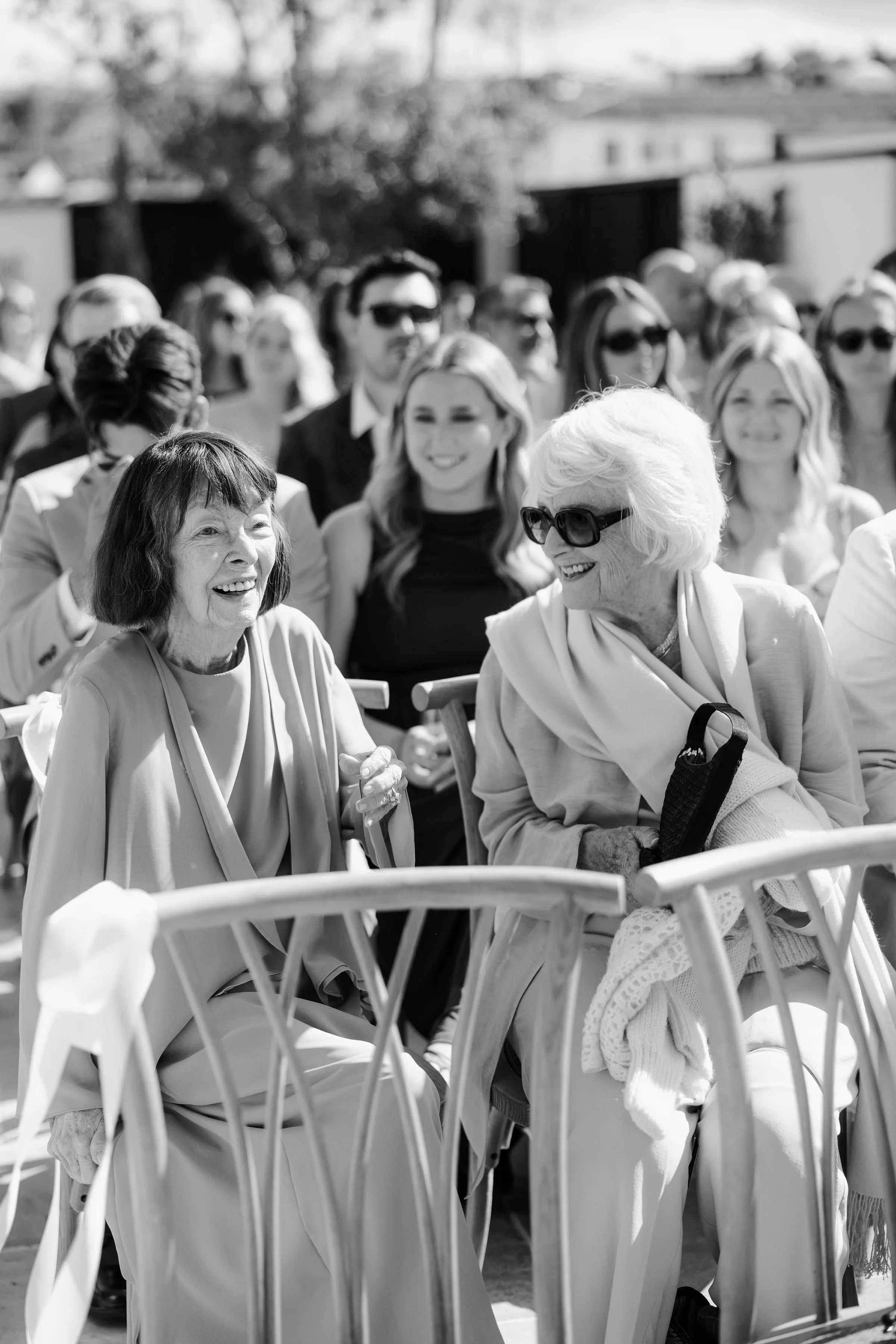A group of elderly women and younger women sitting outdoors, smiling, some wearing sunglasses, with chairs and trees in the background.