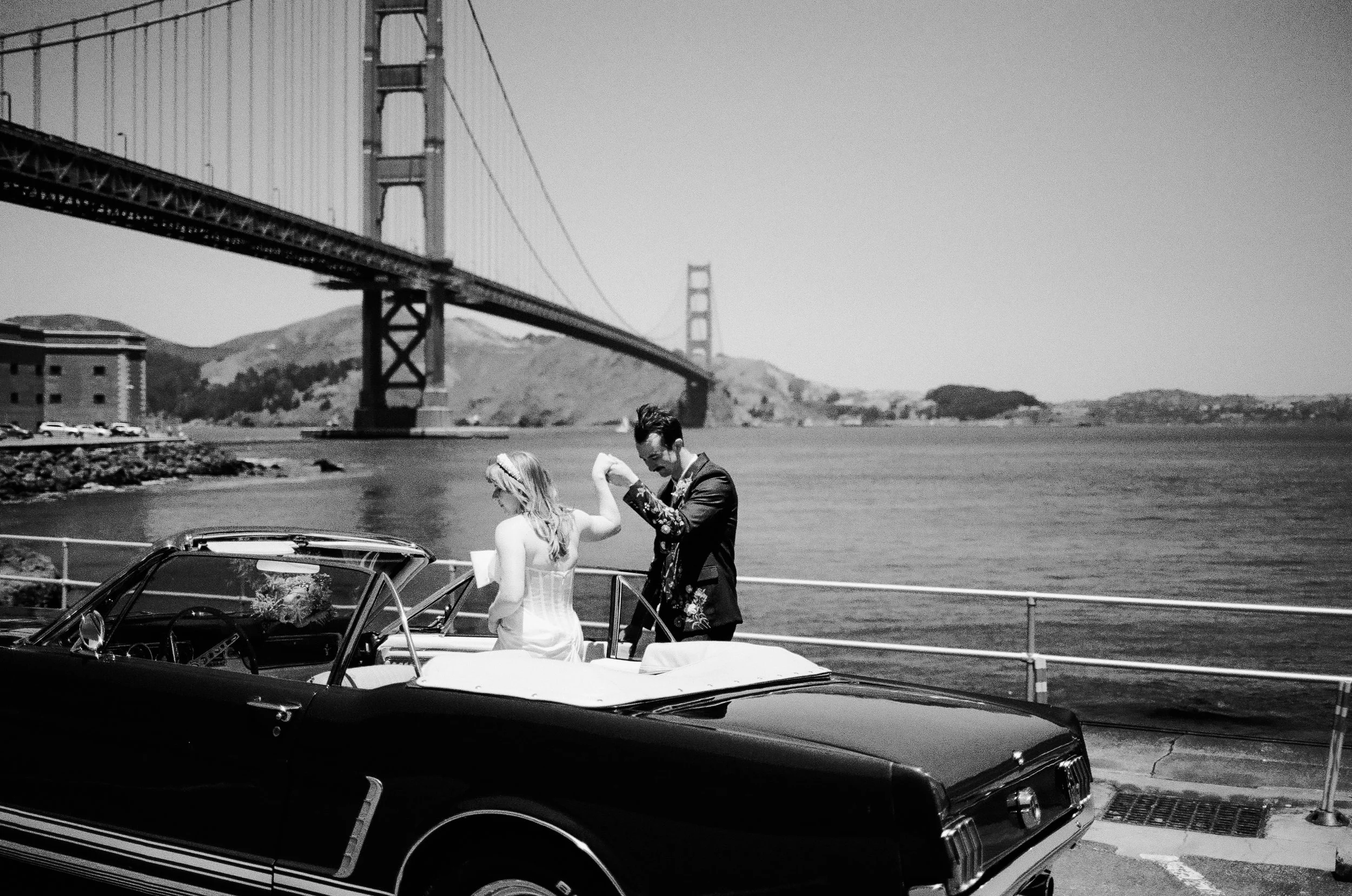 A black-and-white photograph of a bride and groom dancing on a boat docked at a bay with the Golden Gate Bridge in the background. The groom is wearing a floral suit, and the bride is in a strapless wedding dress. A vintage car is parked nearby.