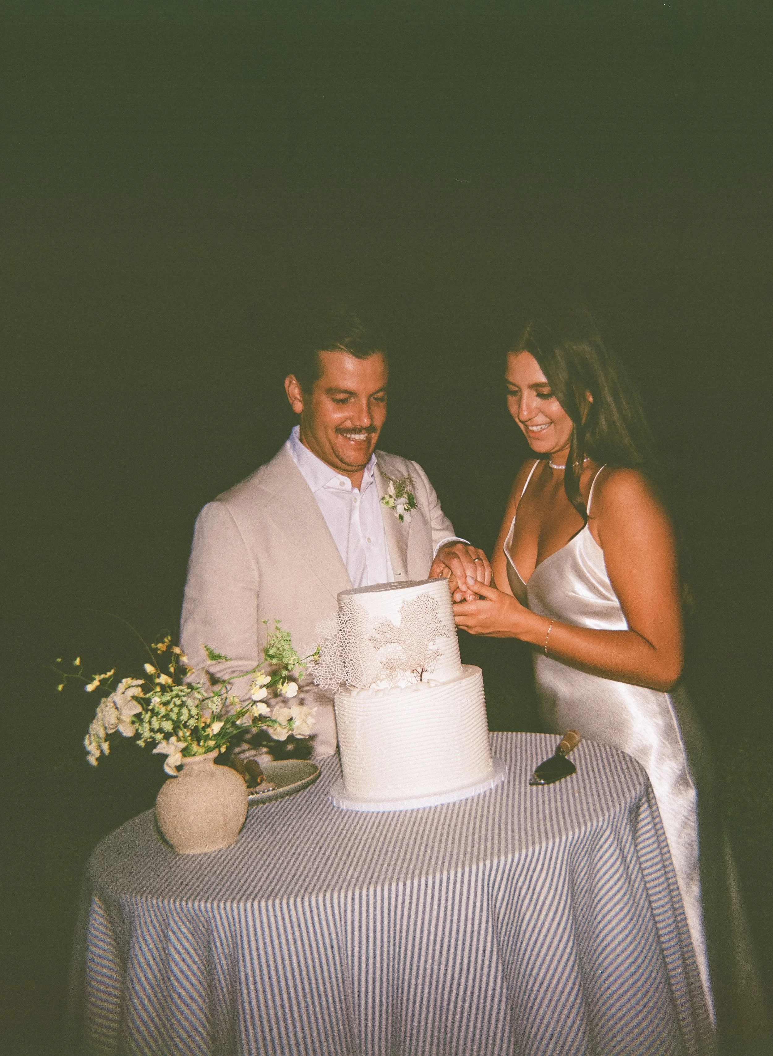 A couple exchanging rings during a wedding ceremony, with a wedding cake and floral arrangements on a table covered with a blue and white striped tablecloth.