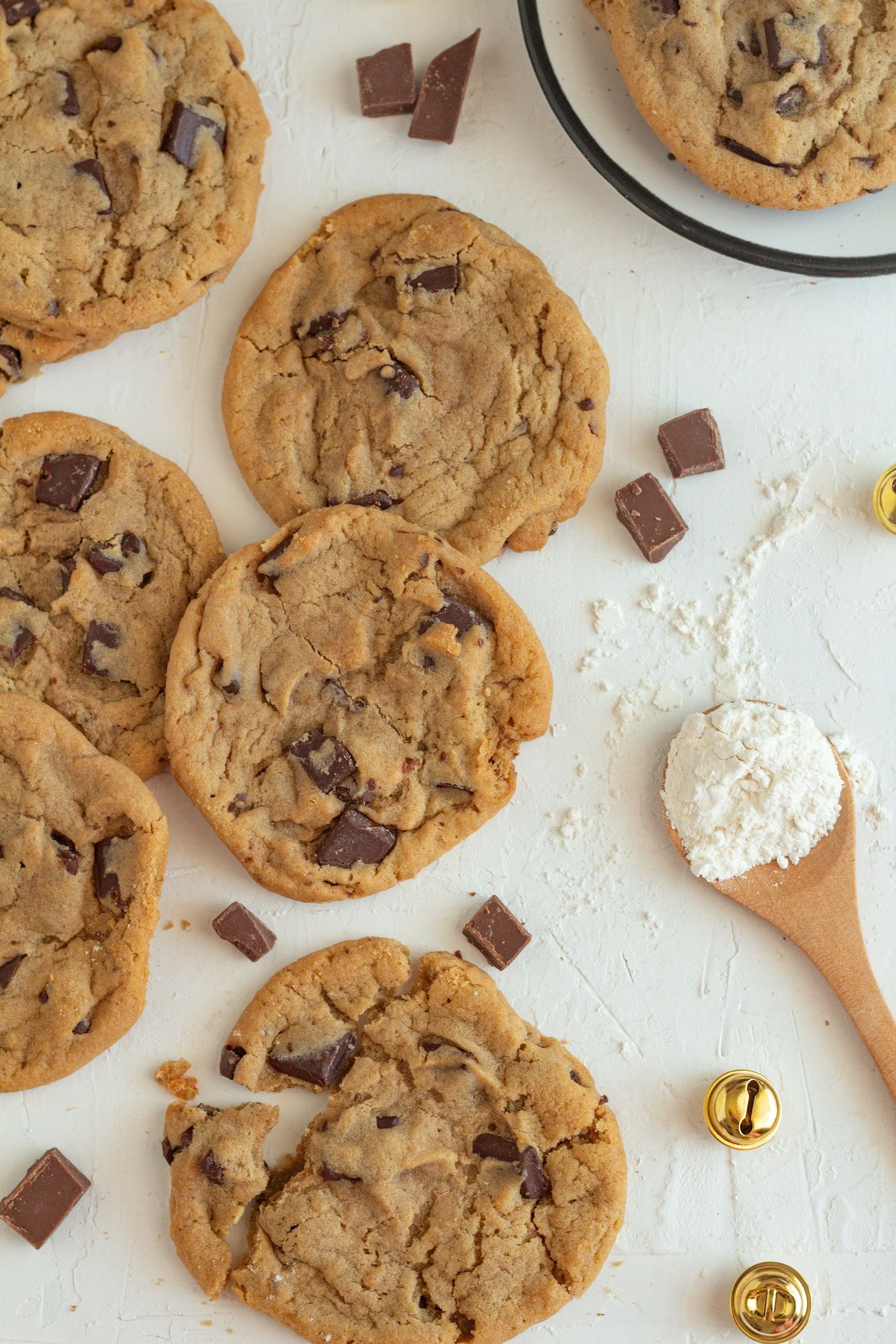 Chocolate chip cookies on a white surface with pieces of chocolate and a cupcake, some sprinkled with powdered sugar, and a small bell.