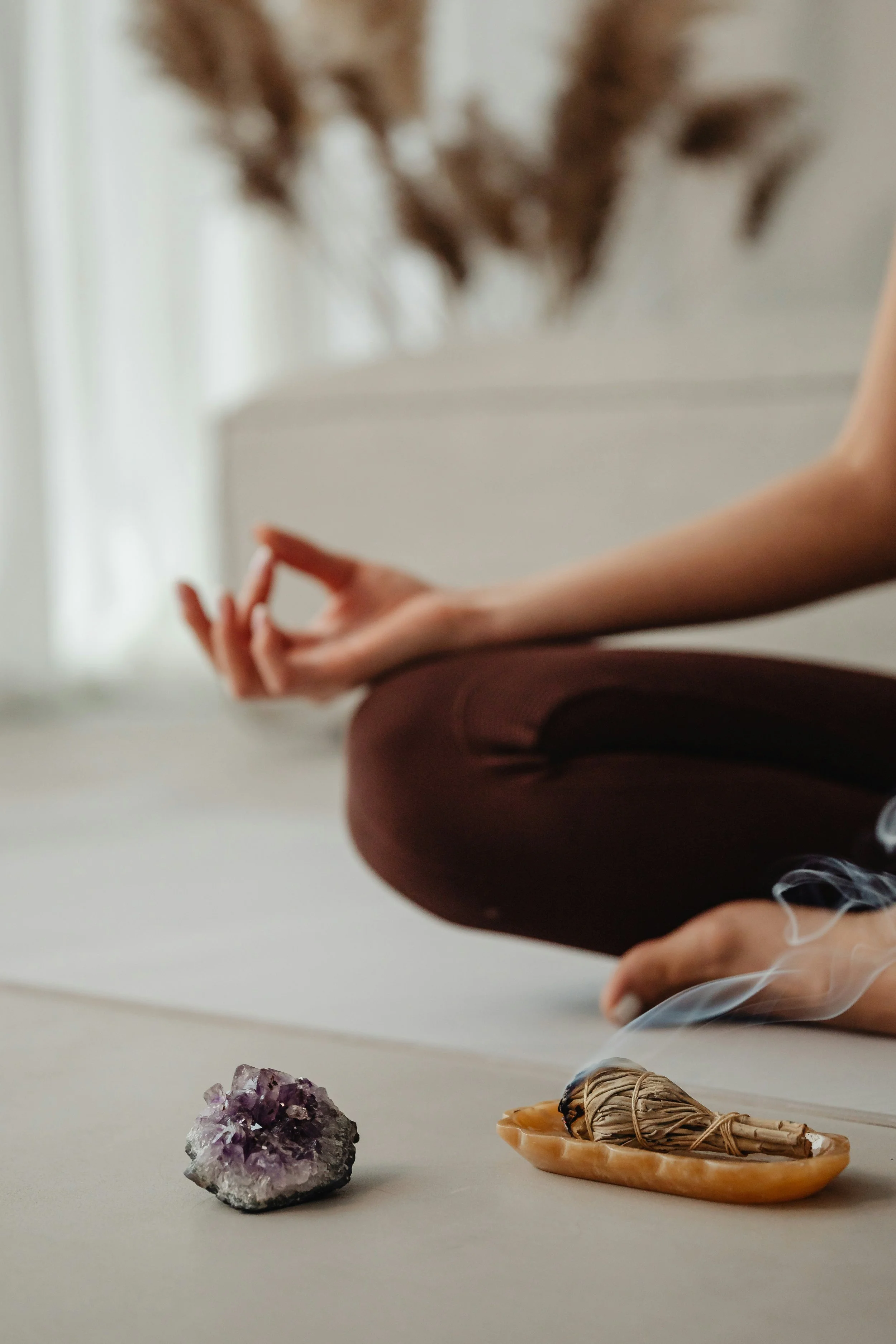 A person sitting cross-legged on the floor with their hands in a meditation mudra, wearing brown pants. In front of them are crystals, a shell with sage bundle, and a piece of amethyst. The background shows a blurred, softly lit space.