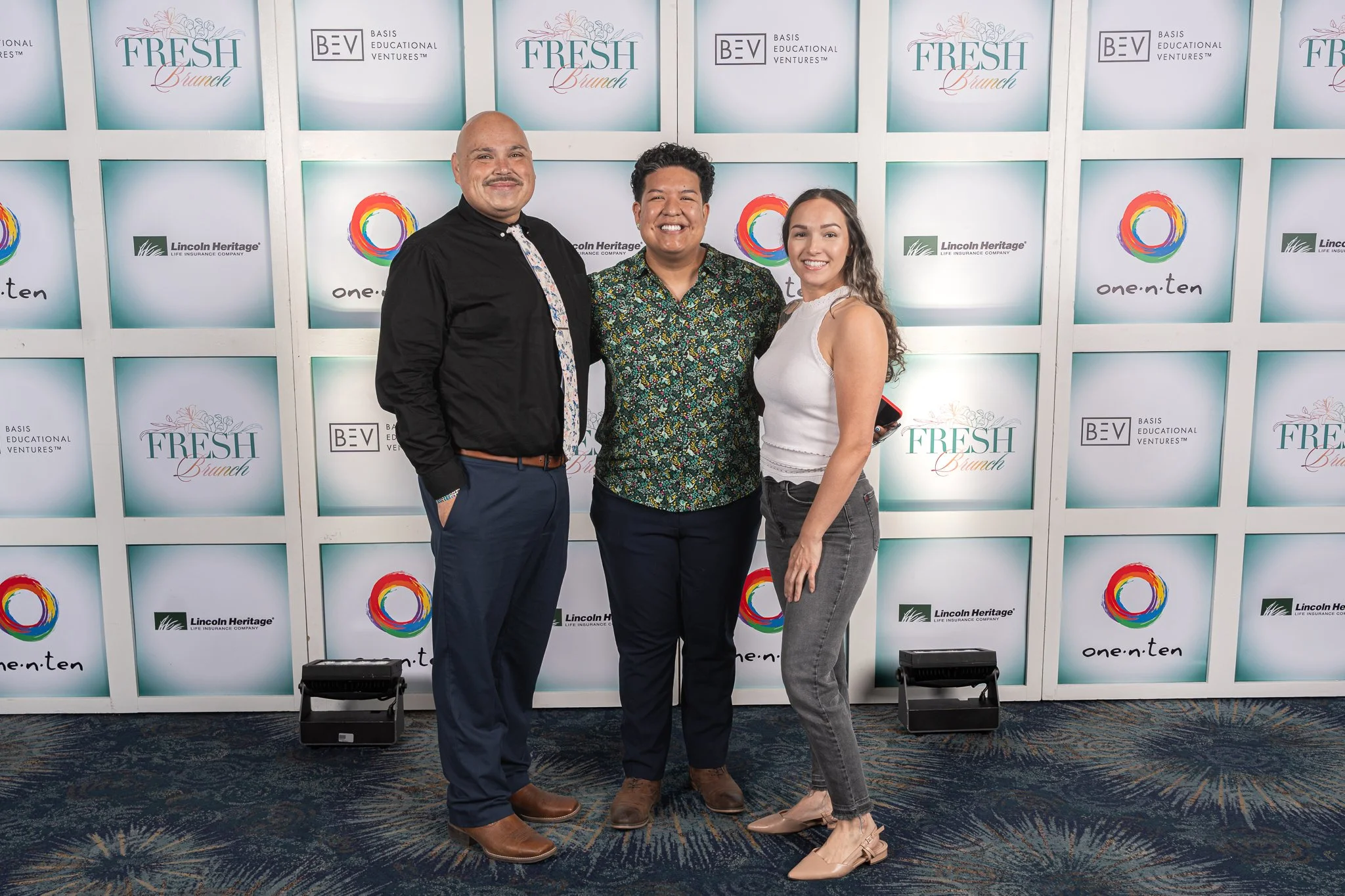 Three people smiling at a conference, standing in front of a backdrop with logos for 'FRESH Brunch,' 'BASIS Educational Ventures,' 'Lincoln Heritage,' and 'one·on·ten.'