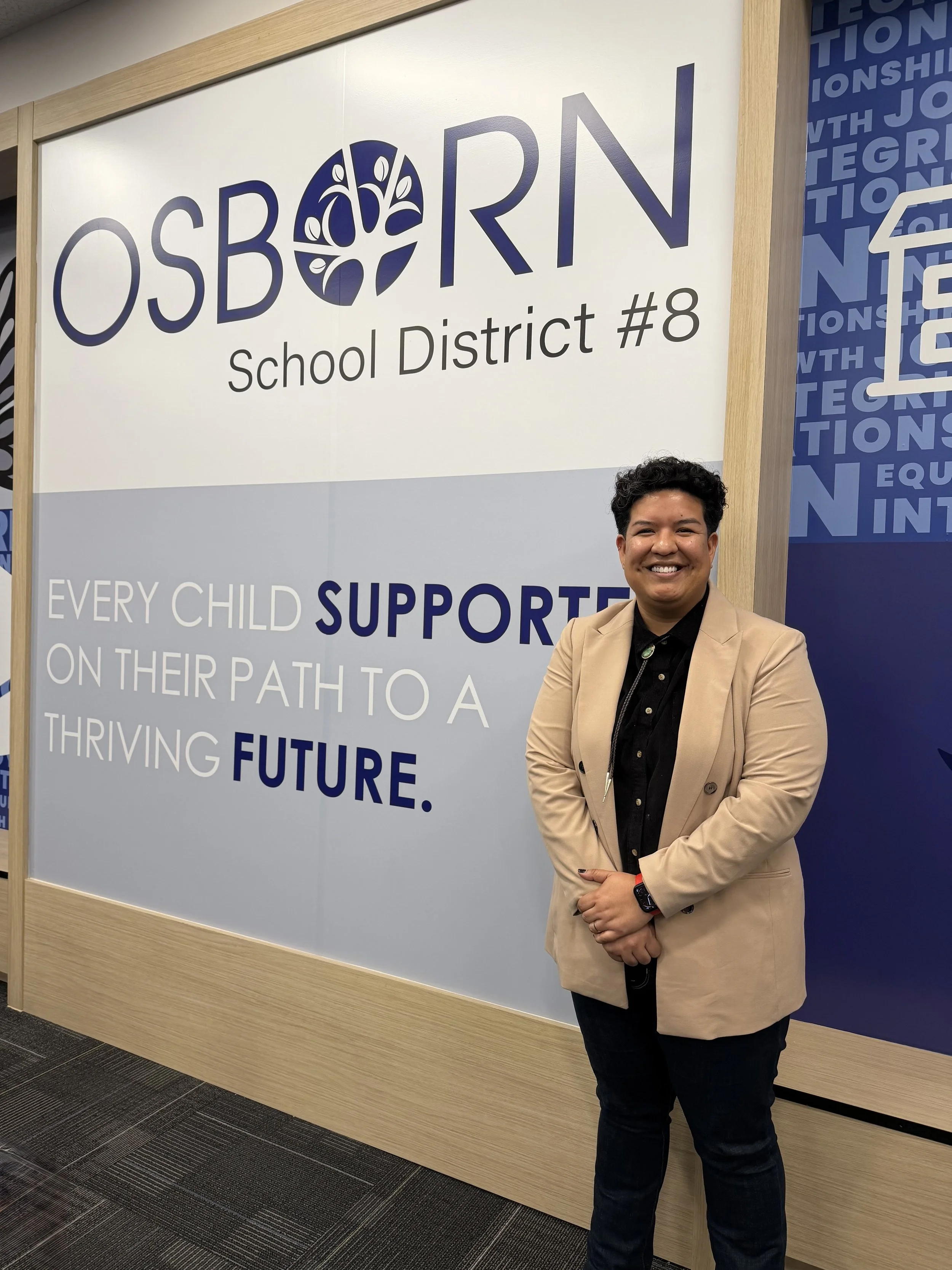 Person in a beige blazer and black shirt standing in front of a large school district sign that reads 'OSBORN, School District #8. Every child supported on their path to a thriving future'.
