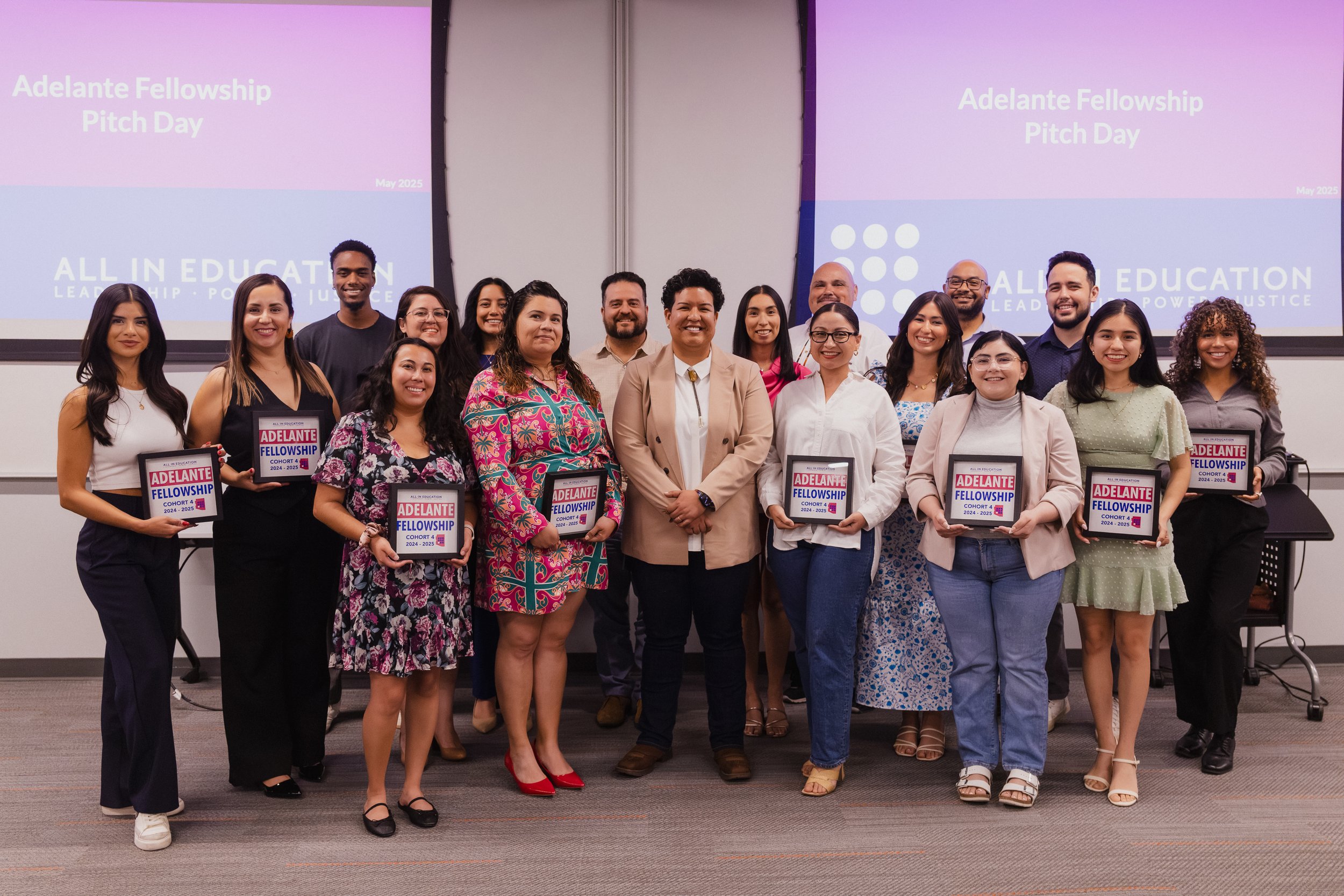 Group of diverse people at an event called Adelante Fellowship Pitch Day, holding certificates, in a conference room with two large screens displaying the event name. The group is smiling and dressed in business casual attire.