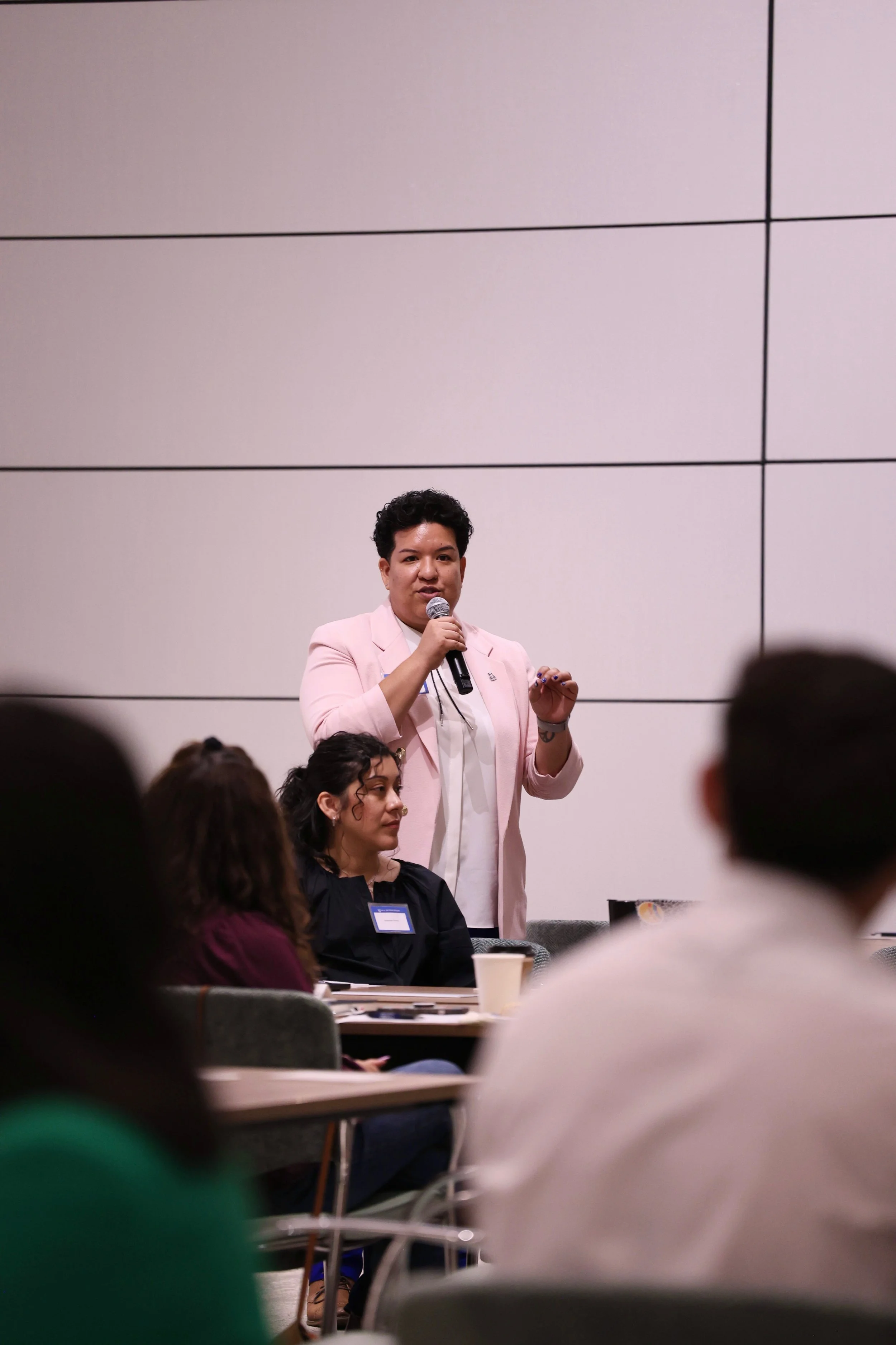 A woman in a pink blazer speaking into a microphone at a conference or seminar, with audience members seated around her.