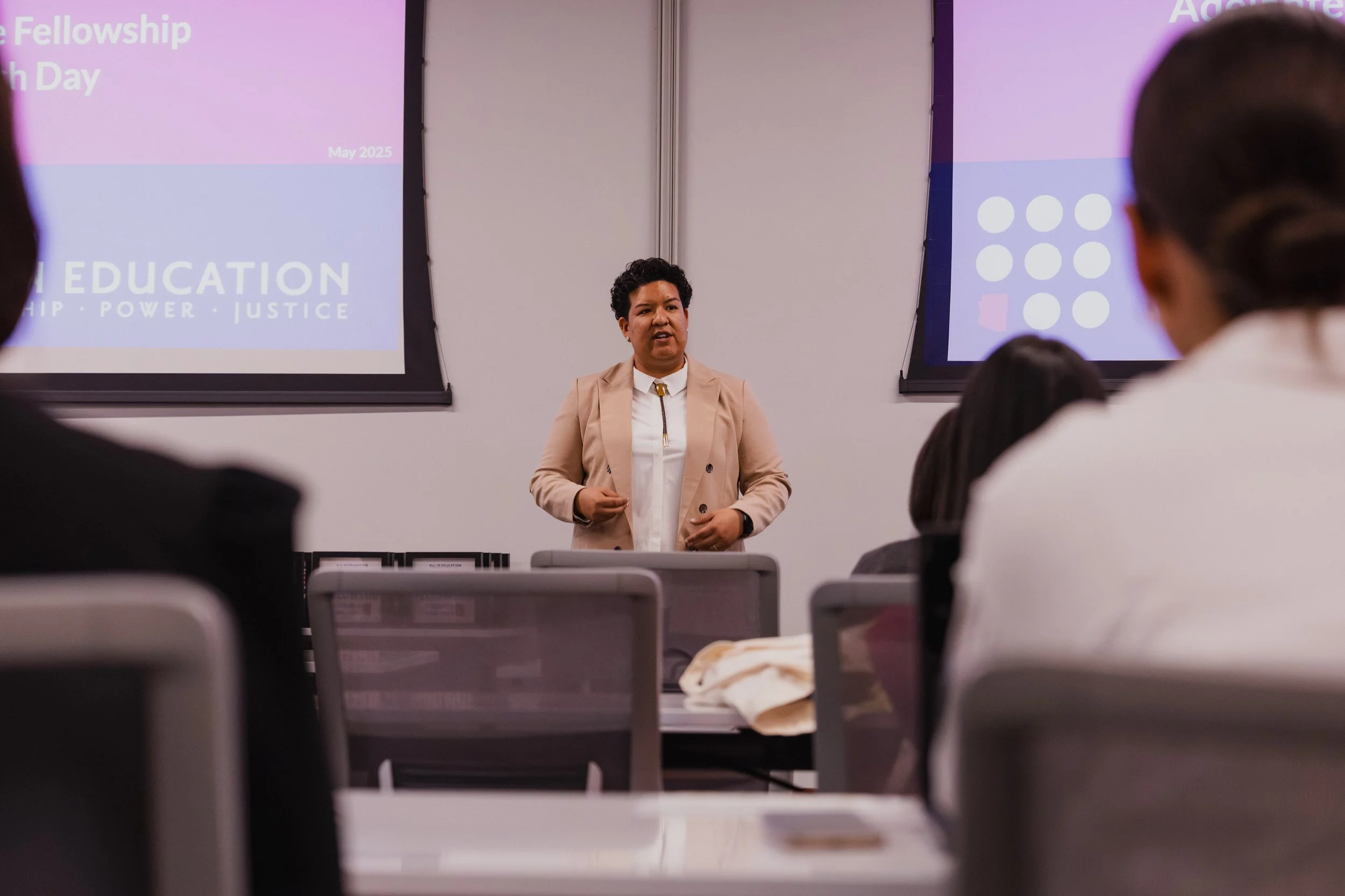 A woman is standing and speaking in front of a classroom with several students seated, with two large presentation screens behind her showing slides related to education and rights, indicating a training or seminar session.