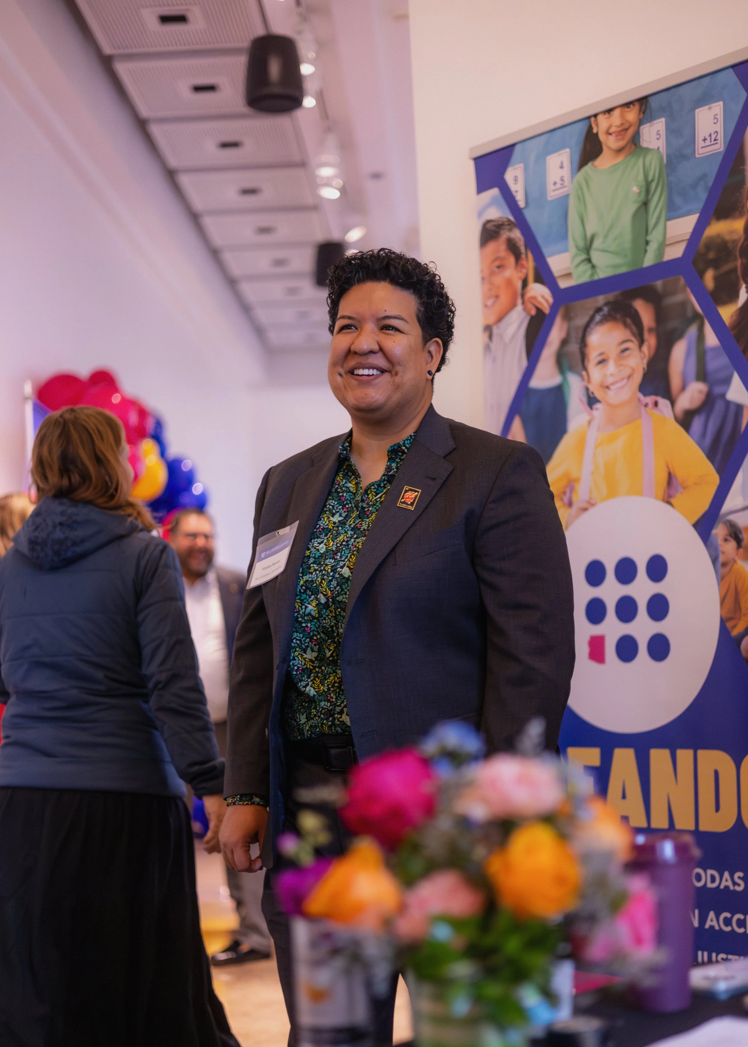 A person with short curly dark hair, wearing a dark blazer and a patterned shirt, smiling at an indoor event with balloons and a colorful banner in the background.