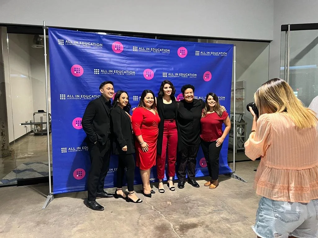 Group of seven people posing for a photo in front of a blue backdrop with the text "All in Education" and pink dot patterns, inside a modern indoor space. One woman on the right is taking their picture.