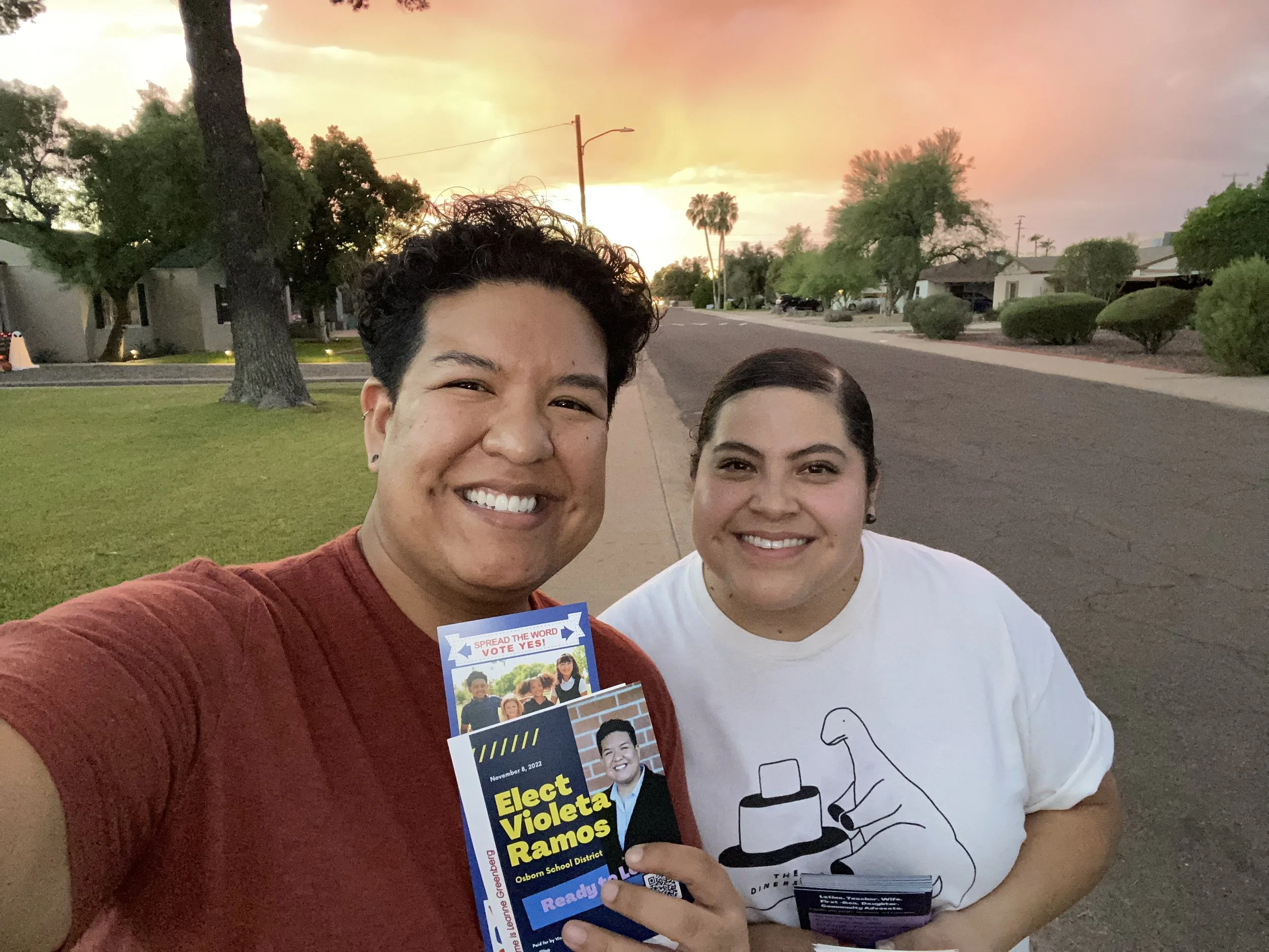 Two smiling people taking a selfie outdoors during sunset, one holding a campaign flyer for Violeta Ramos, with trees and a quiet suburban street in the background.