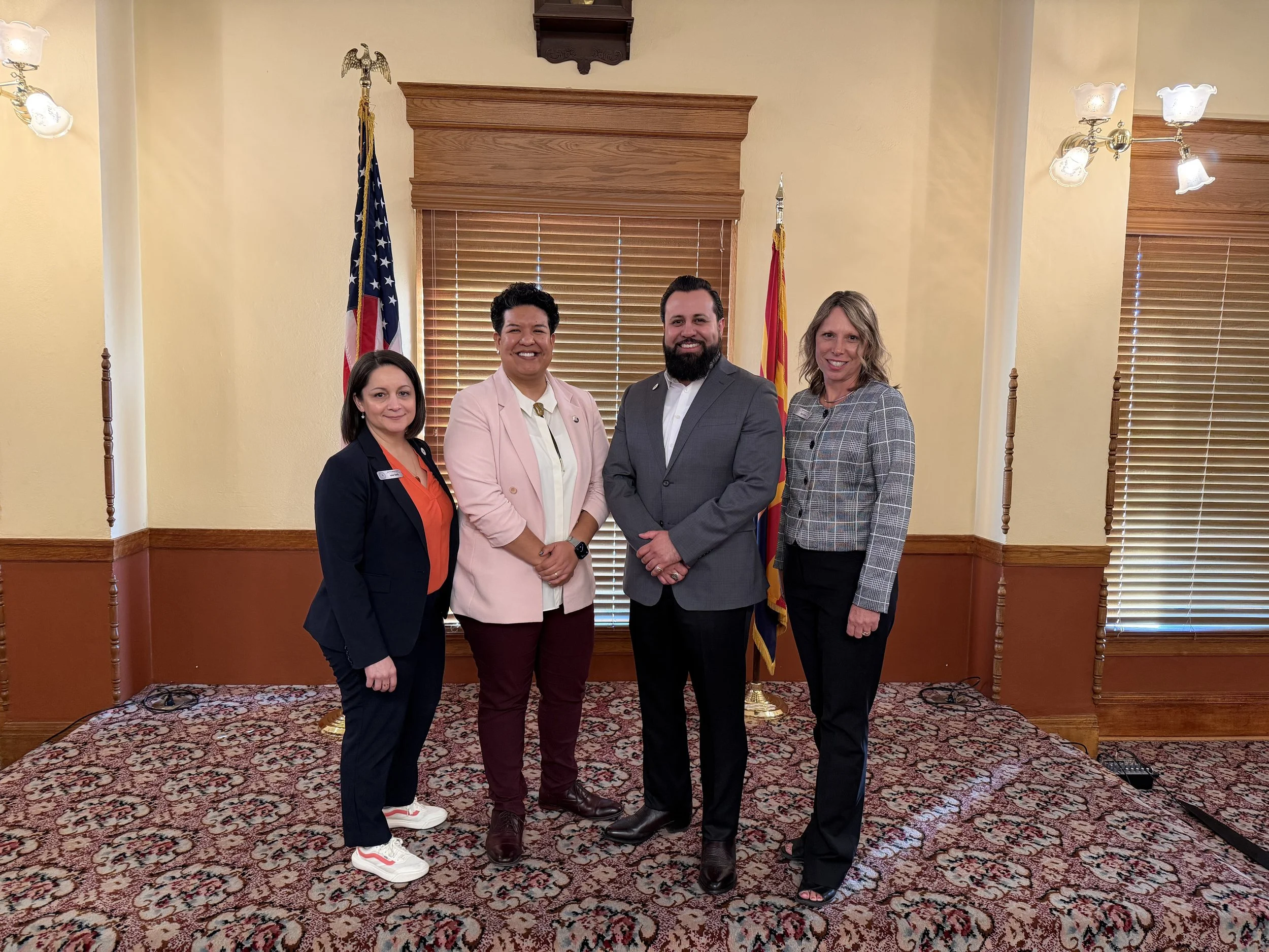 Group of four people standing in front of American and state flags in a formal room with wooden blinds and patterned carpet, smiling at the camera.