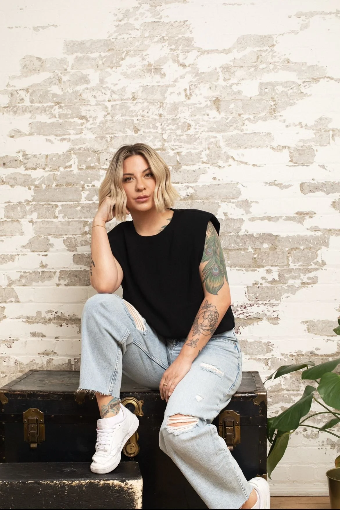 A young woman with blonde wavy hair and tattoos on her arm, sitting on an old black trunk, wearing a black sleeveless top, ripped light blue jeans, and white sneakers, against a white brick wall.