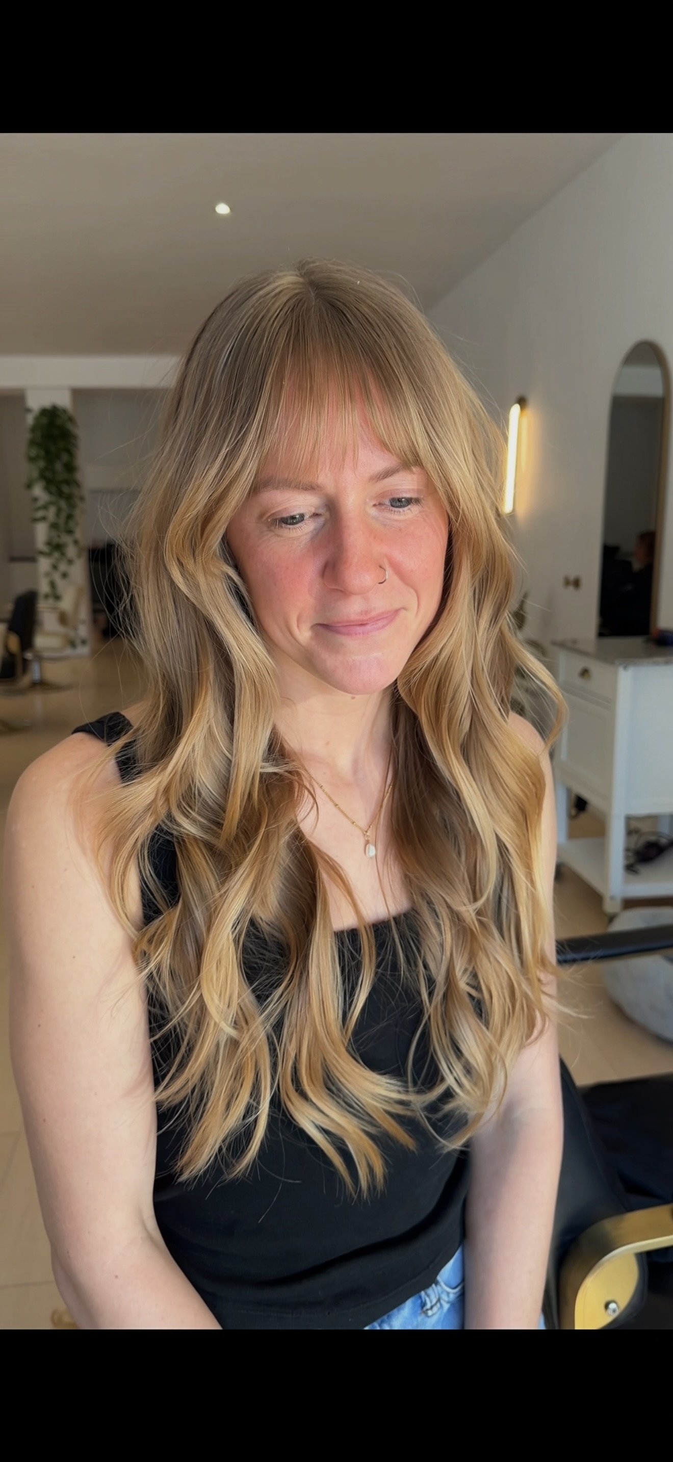 A woman with long, wavy blonde hair wearing a black sleeveless top, a gold necklace, and a nose ring, standing inside a modern, well-lit room with white walls and furniture.