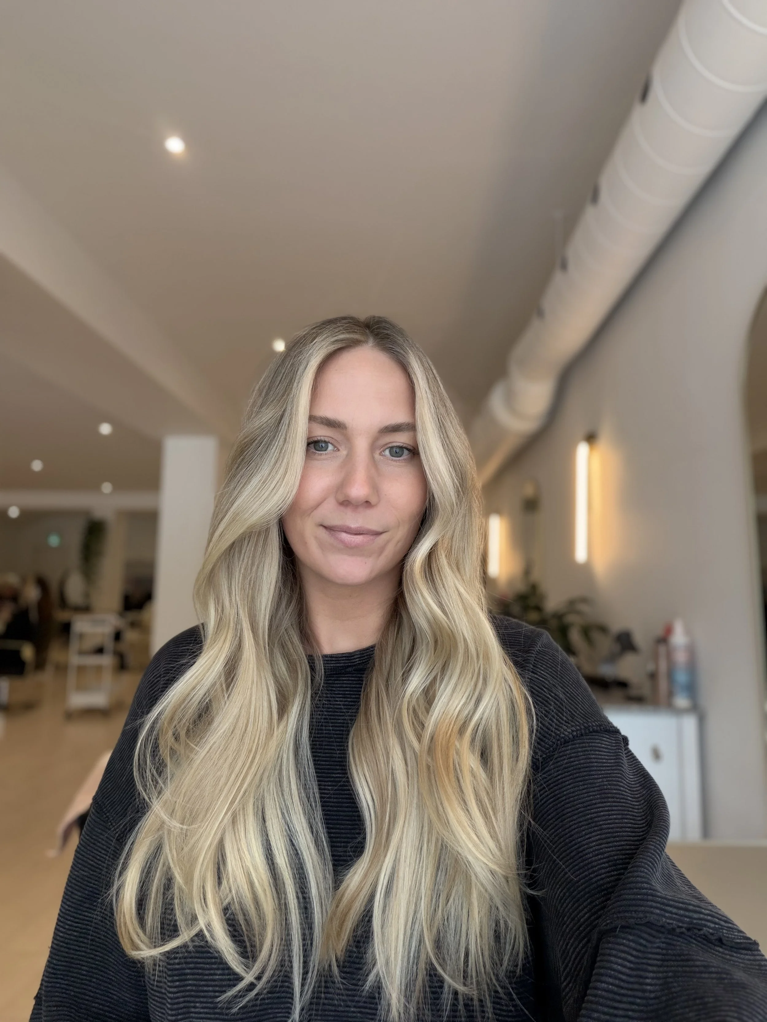 A young woman with long blonde wavy hair and light blue eyes, wearing a black long-sleeve shirt, smiling in a modern indoor setting with beige walls, white ceiling, and warm lighting.