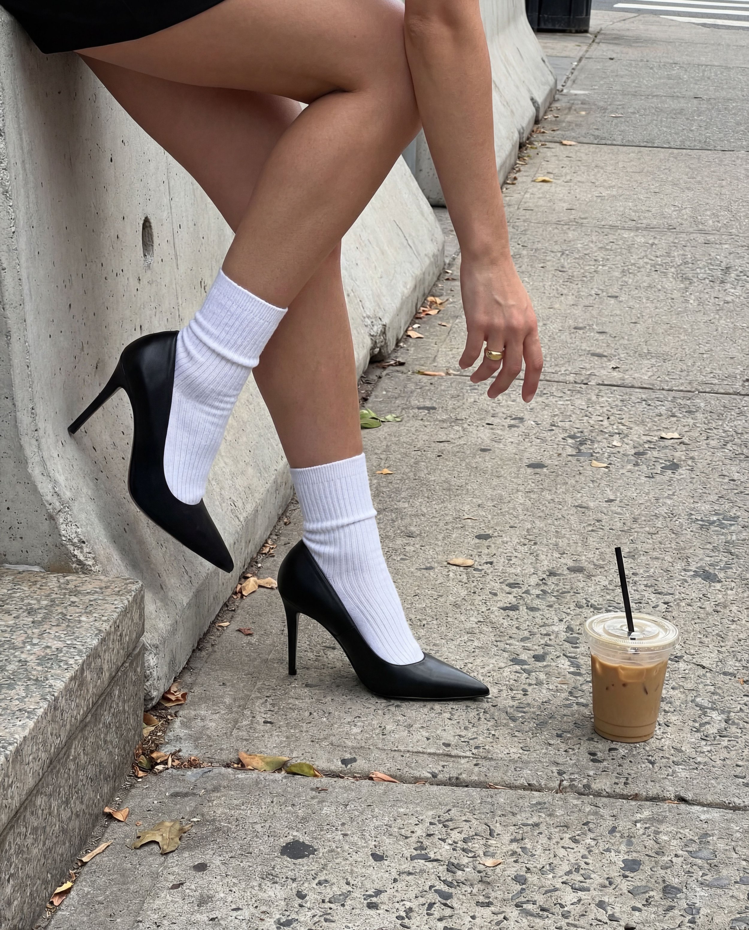 Close-up of a person's legs, feet, and hand as they sit on a concrete curb sidewalk. The person is wearing black high heels, white socks, and a ring on their finger. There is a plastic cup with iced coffee on the ground nearby. The background shows a sidewalk and a street with some fallen leaves.