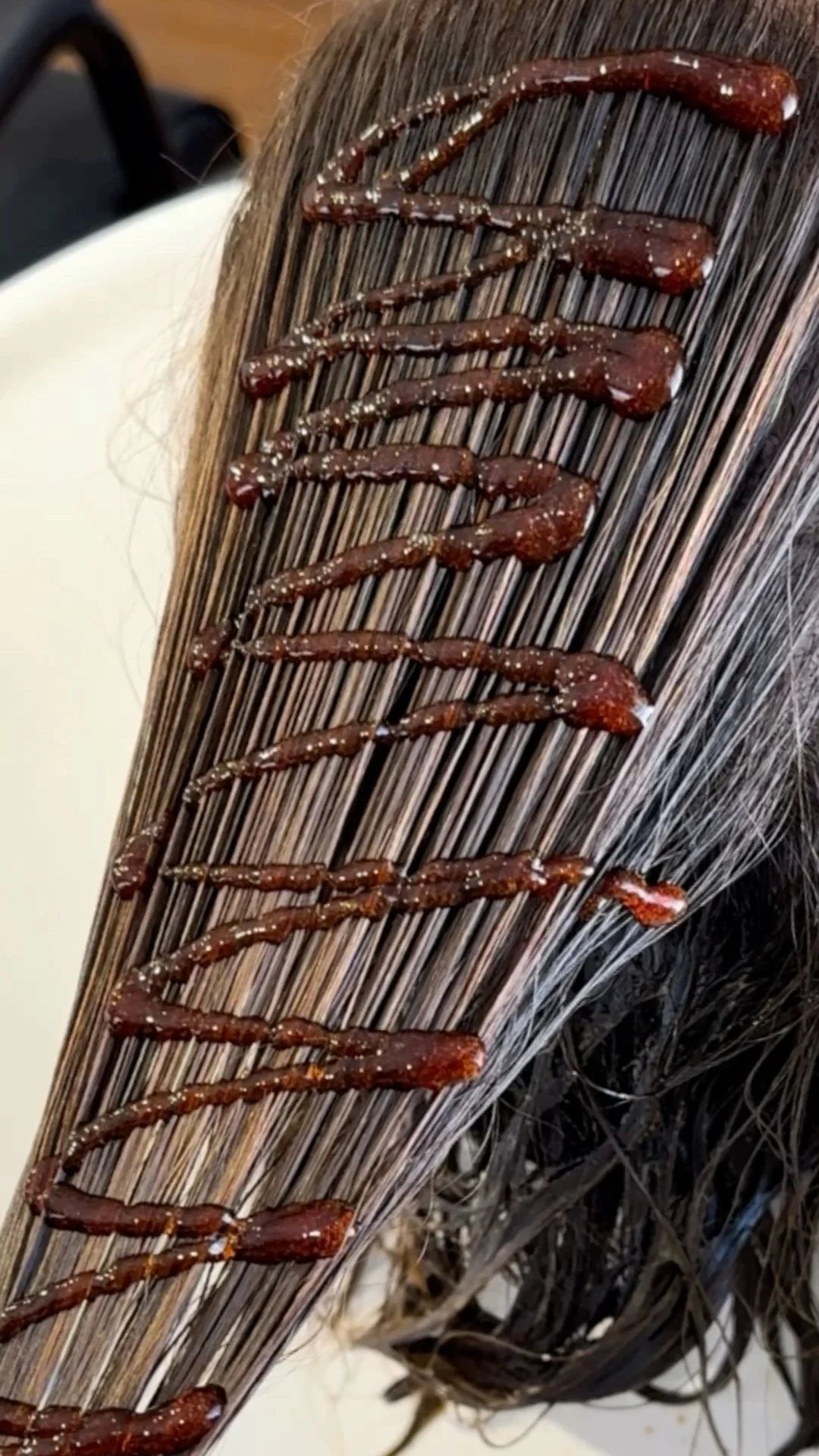 Close-up of a hair strand with a brown hair dye or treatment applied, decorated with a zigzag pattern of a darker, gel-like substance with white specks.