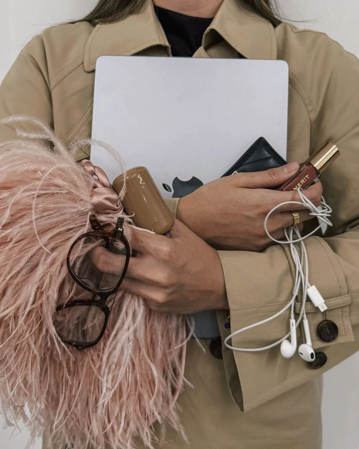 Person holding a MacBook, a perfume bottle branded 'MERCIT', a pair of sunglasses, a beige fluffy clutch, and earphones. The person is wearing a beige trench coat.