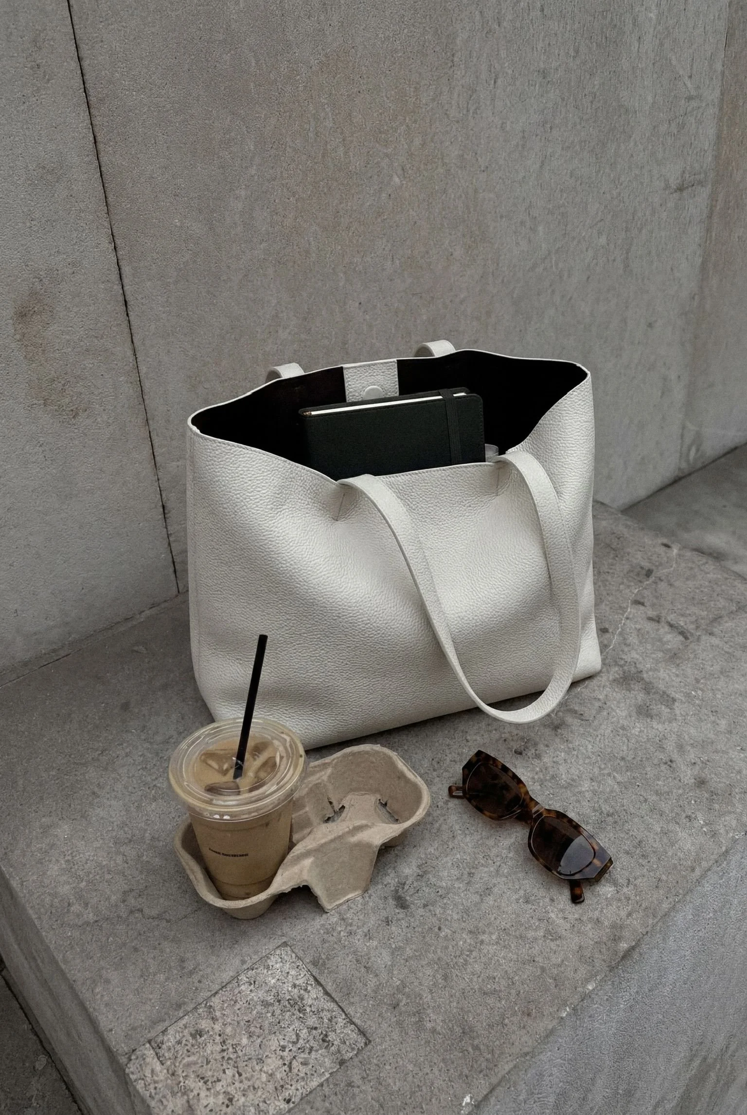 A white textured tote bag with two handles and an open top, placed on a concrete surface against a gray wall, with a black notebook inside. Nearby on the ground, there is a plastic cup of iced coffee with a straw, a brown cardboard drink holder, and a pair of tortoise shell sunglasses.