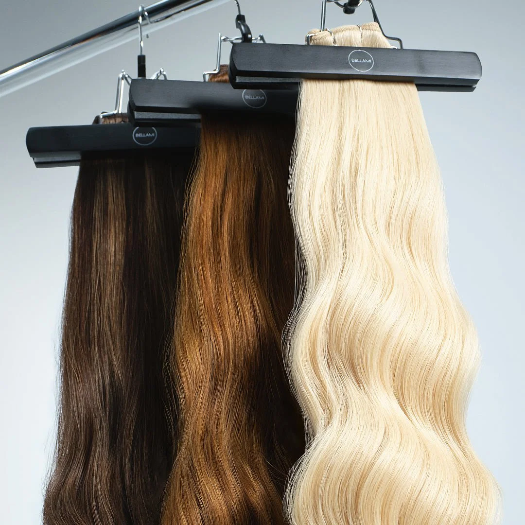 Three hair extensions in shades of dark brown, auburn, and platinum blonde hanging on black clips from a metal rod.