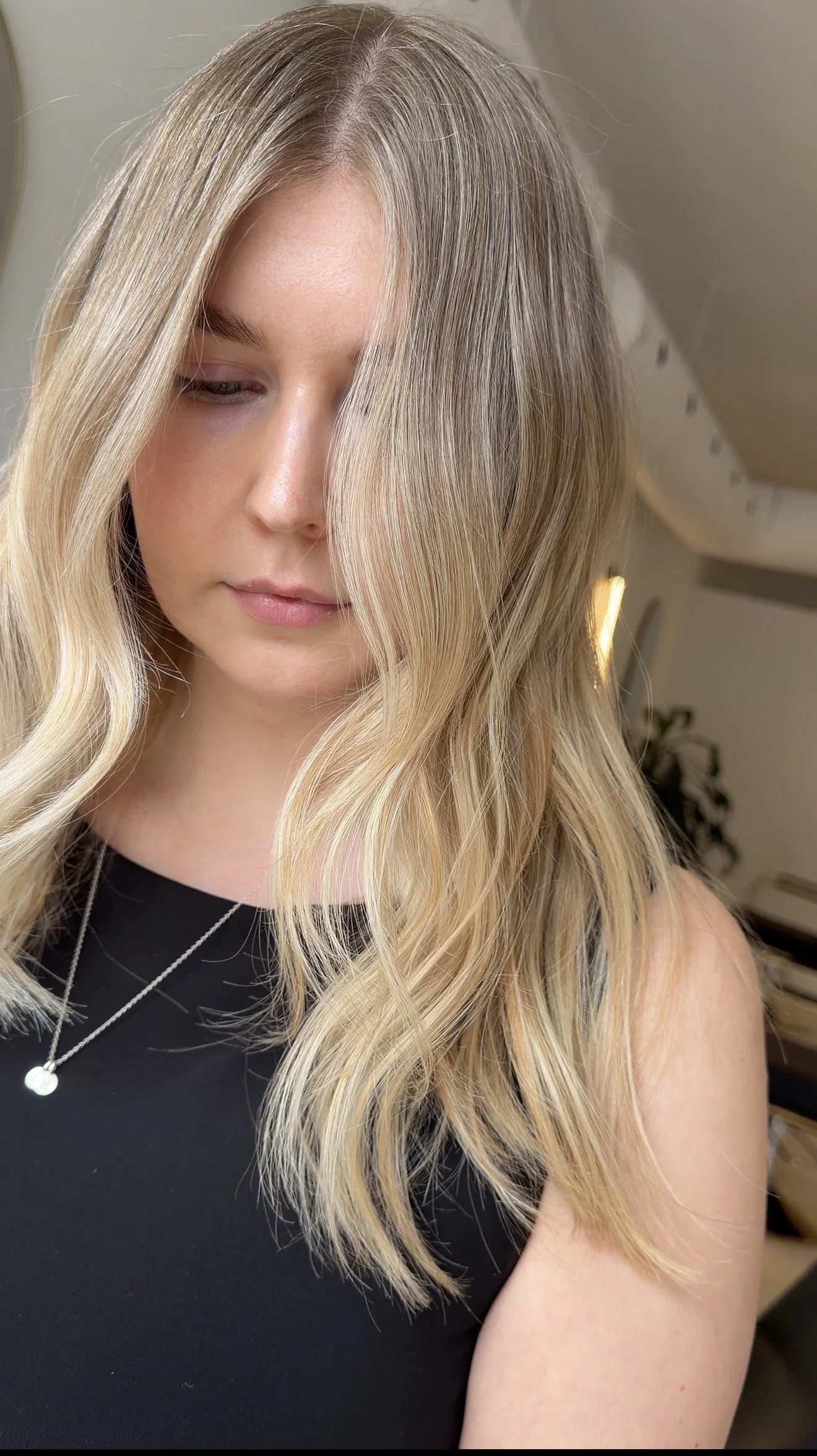 Close-up of a woman with long, wavy blonde hair covering part of her face, wearing a black top and layered necklaces, indoor setting with soft lighting.