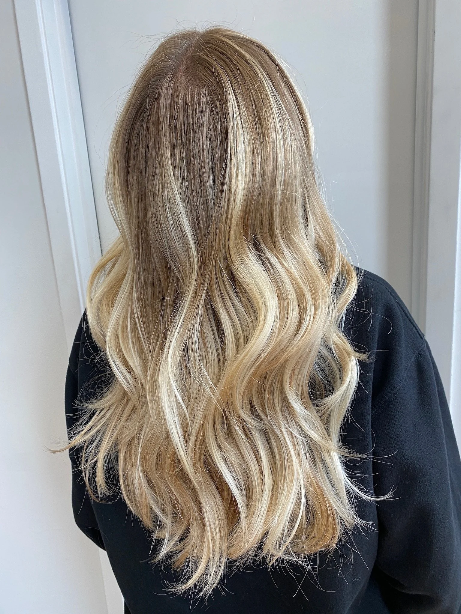 Close-up of a person's head with long, wavy blonde hair, wearing a black shirt, standing near a white wall.