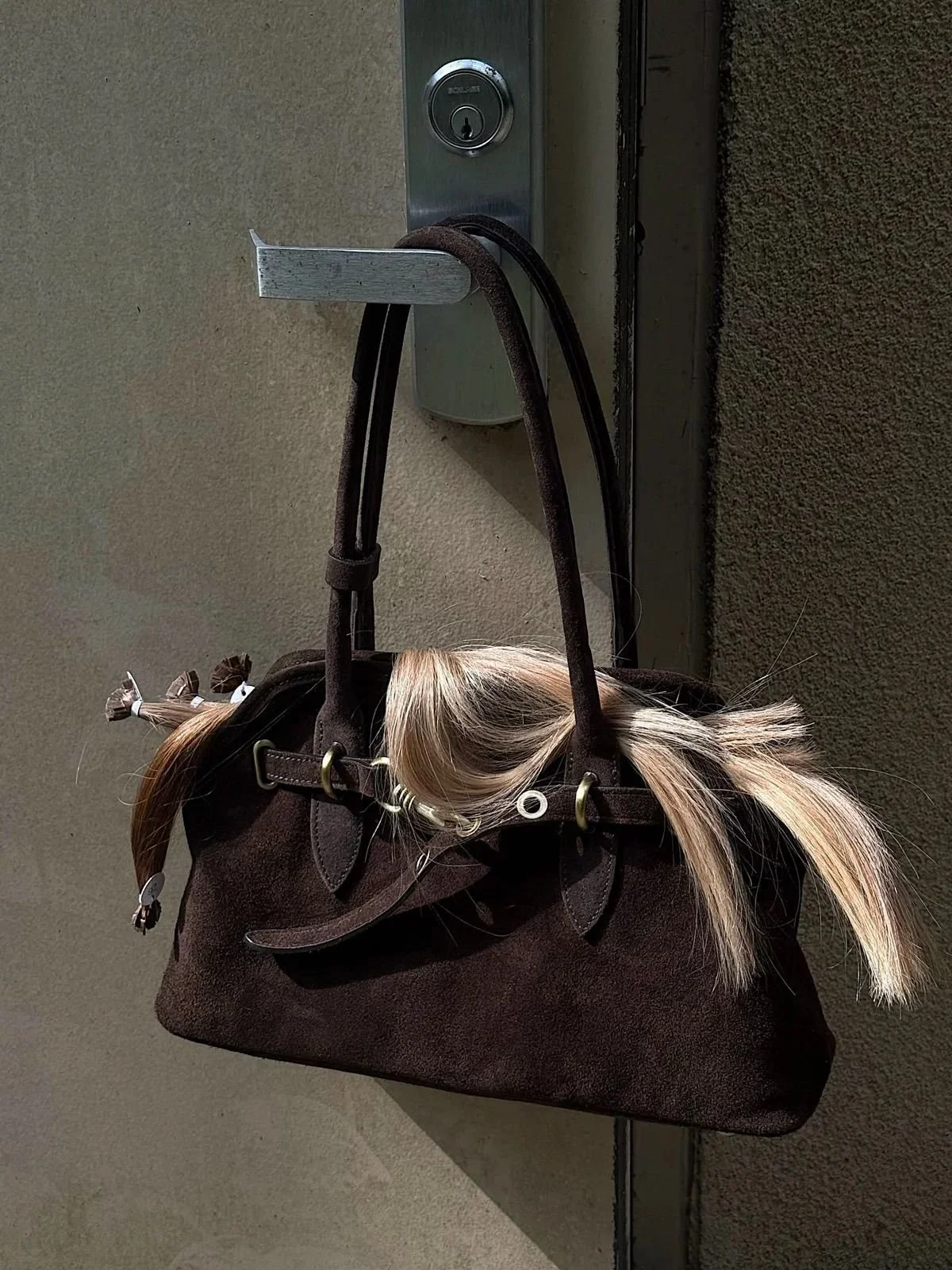 A brown handbag with a blonde hair extension attached, hanging on a door handle next to a keyhole.
