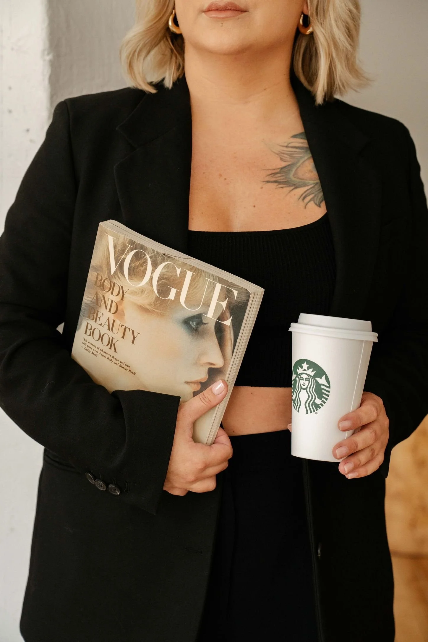 A woman with blonde hair holding a Vogue magazine and a Starbucks cup, wearing a black blazer and black crop top, standing against a plain background.