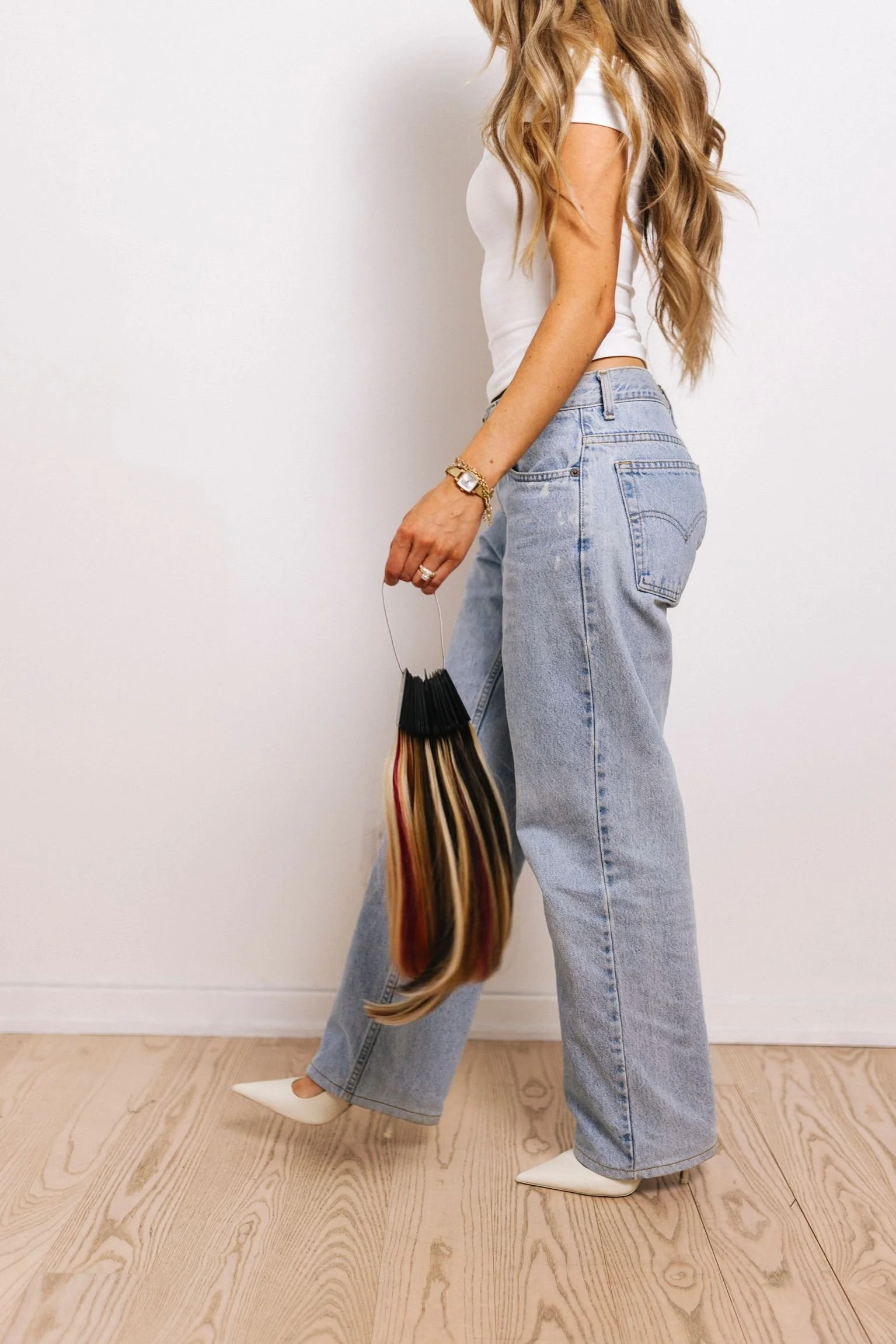 A woman with long wavy blonde hair wearing a white fitted t-shirt and high-waisted light blue jeans, holding a multicolored shopping bag, standing indoors against a white wall and light wooden floor.