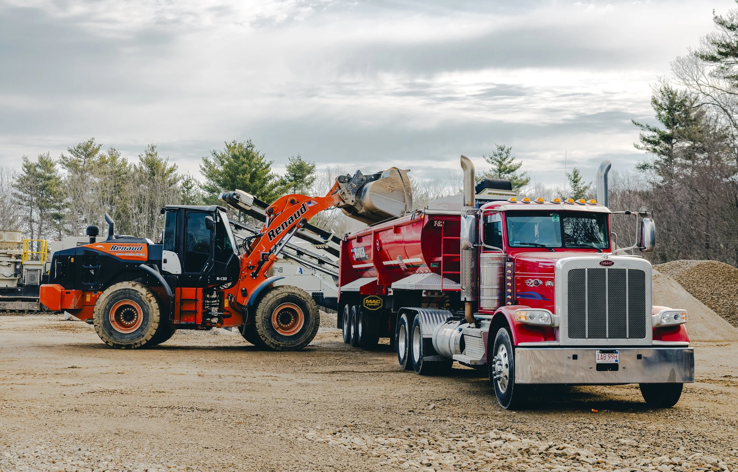 A red semi-truck with chrome exhaust stacks parked on a dirt lot next to an orange and white backhoe loader with the brand name Renaud. The background includes trees and a cloudy sky.