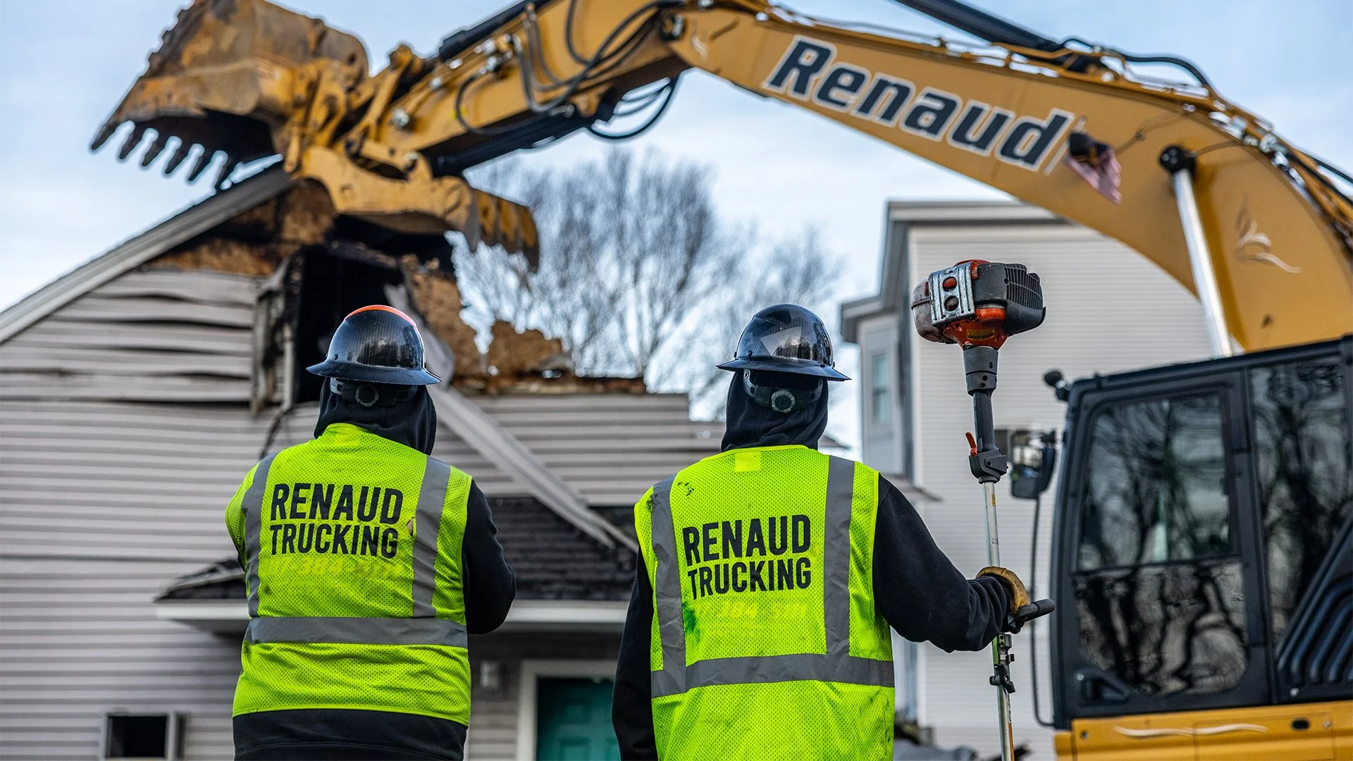 Workers wearing yellow safety vests and helmets operating heavy machinery to demolish a house, with part of the house's roof collapsed.