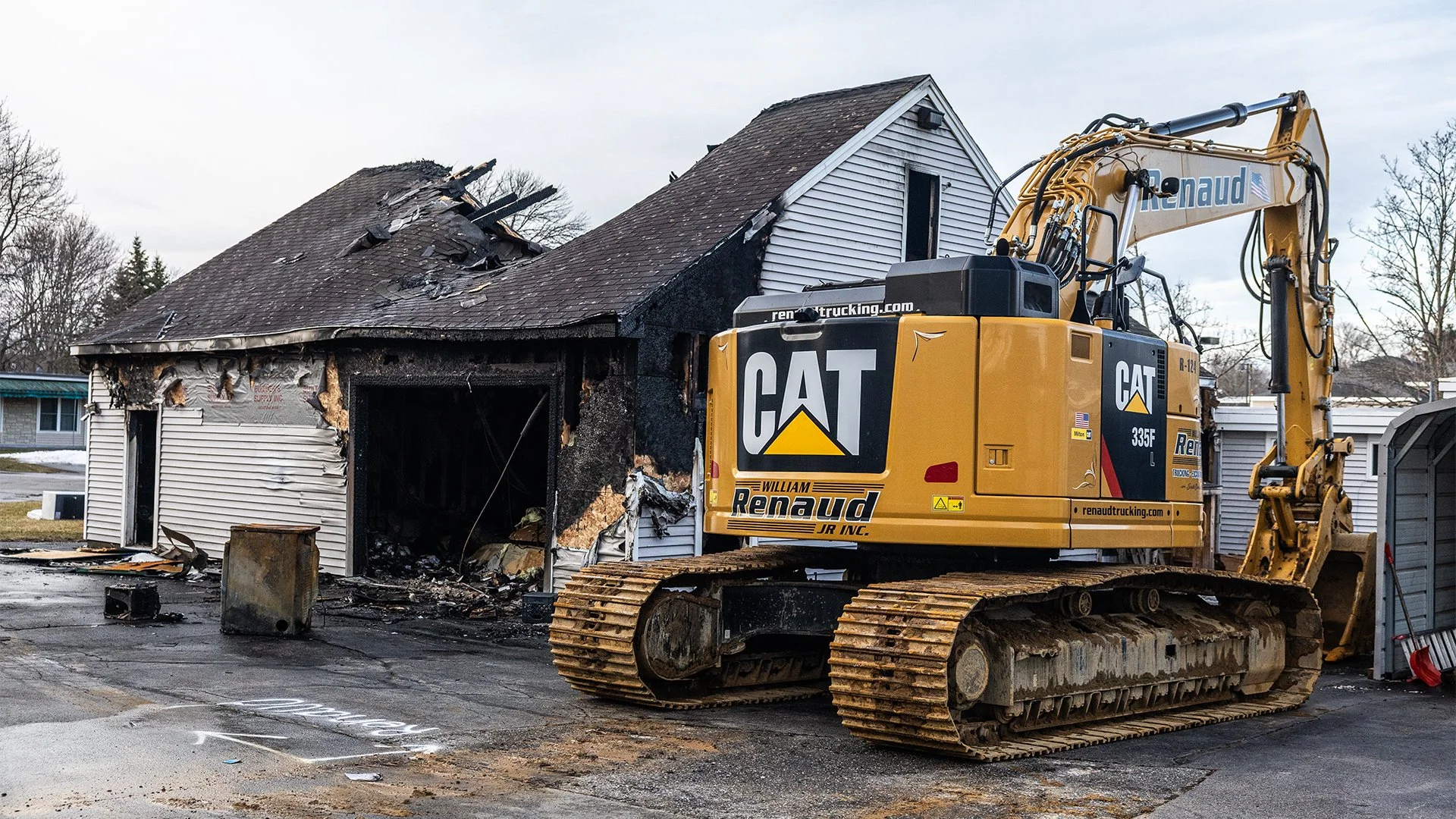 A damaged house with significant fire damage and charring on the exterior walls, with a large yellow Caterpillar excavator nearby, likely used for cleanup after a fire.