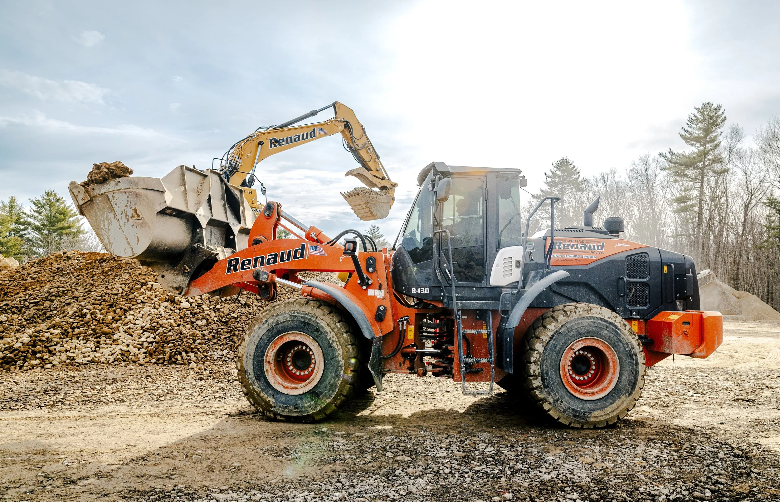 Construction site with an orange and black Renaud tractor and a yellow excavator loading gravel or soil.