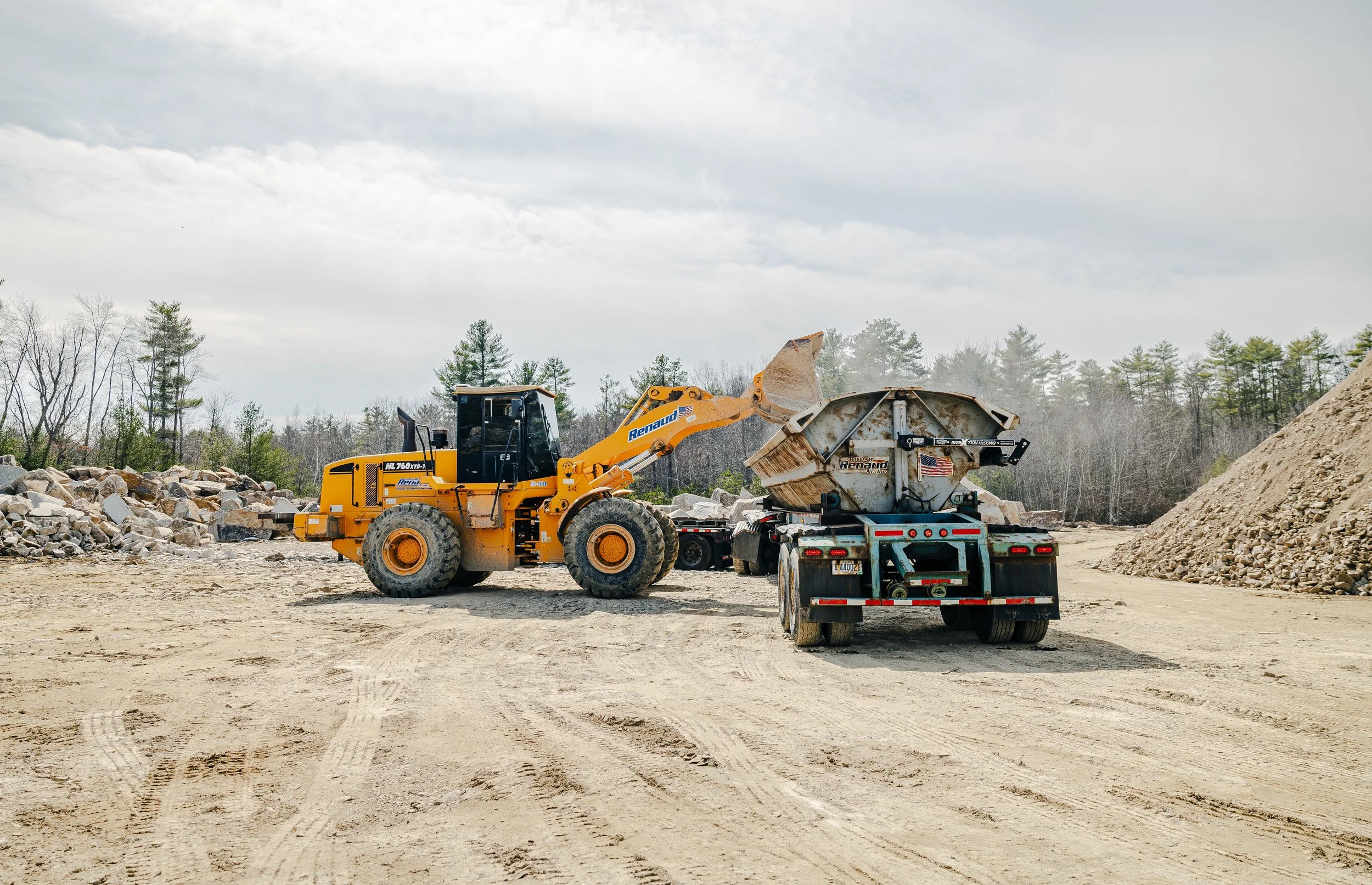 Construction site with a large yellow front loader and a flatbed truck delivering soil or construction material, with dirt and rocks on the ground and trees in the background.