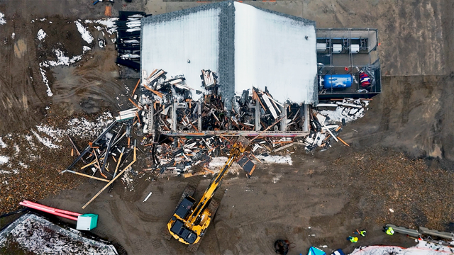 Overhead view of a partially demolished building with debris, a construction excavator, and construction workers on-site.