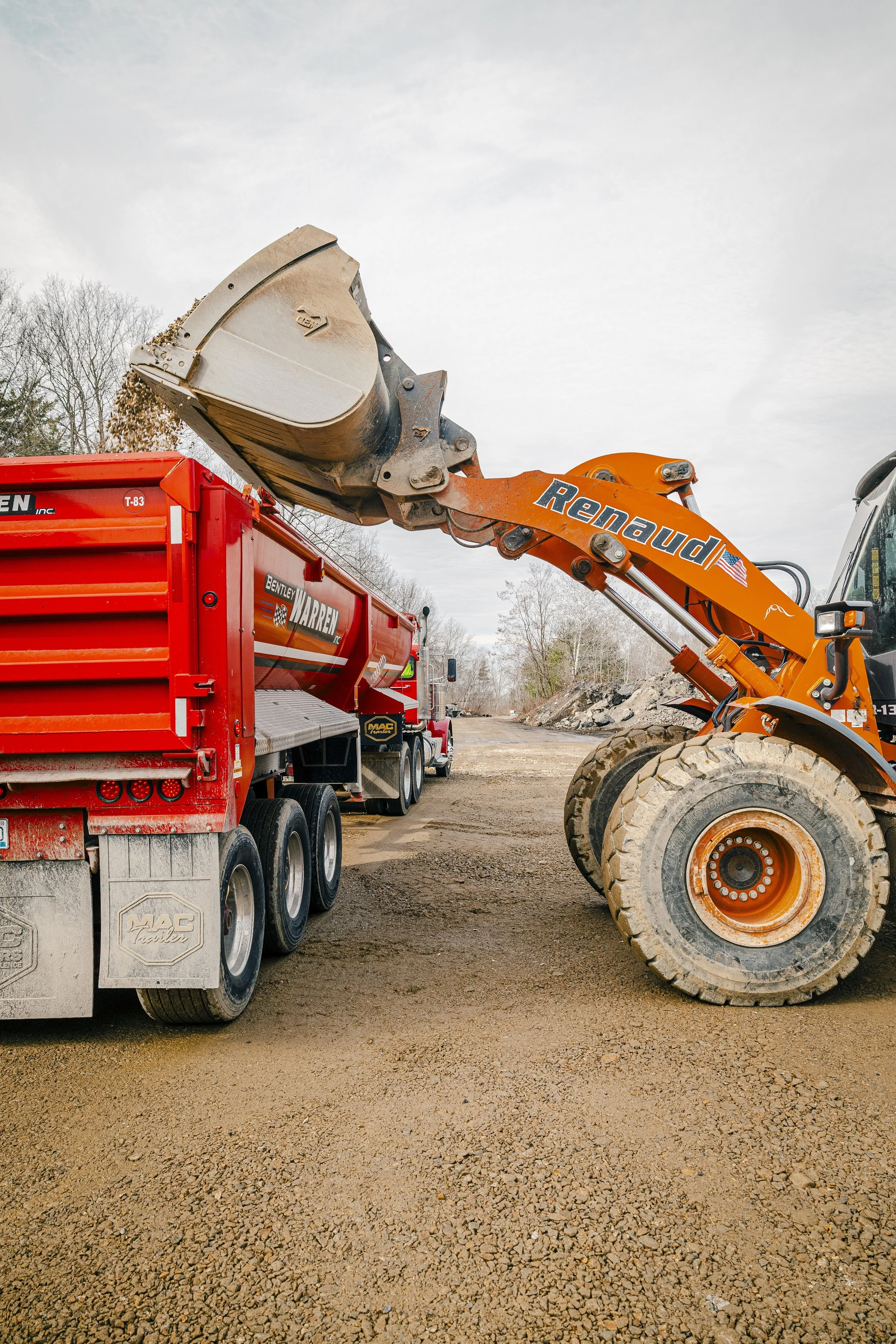 Construction site with a large orange excavator lifting dirt into a red dump truck, gravel ground, cloudy sky, trees in the background.