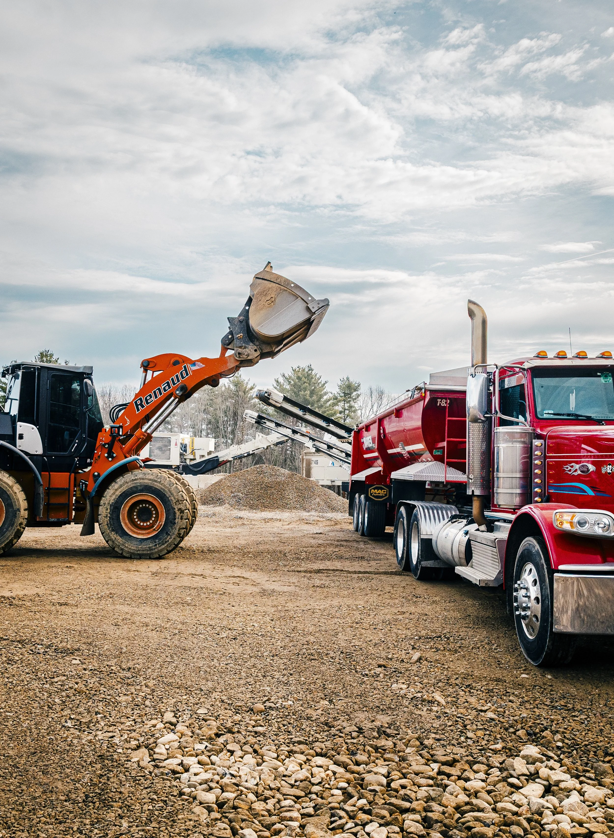 Construction site with a large red dump truck and an orange excavator lifting a scoop of dirt against a cloudy sky.