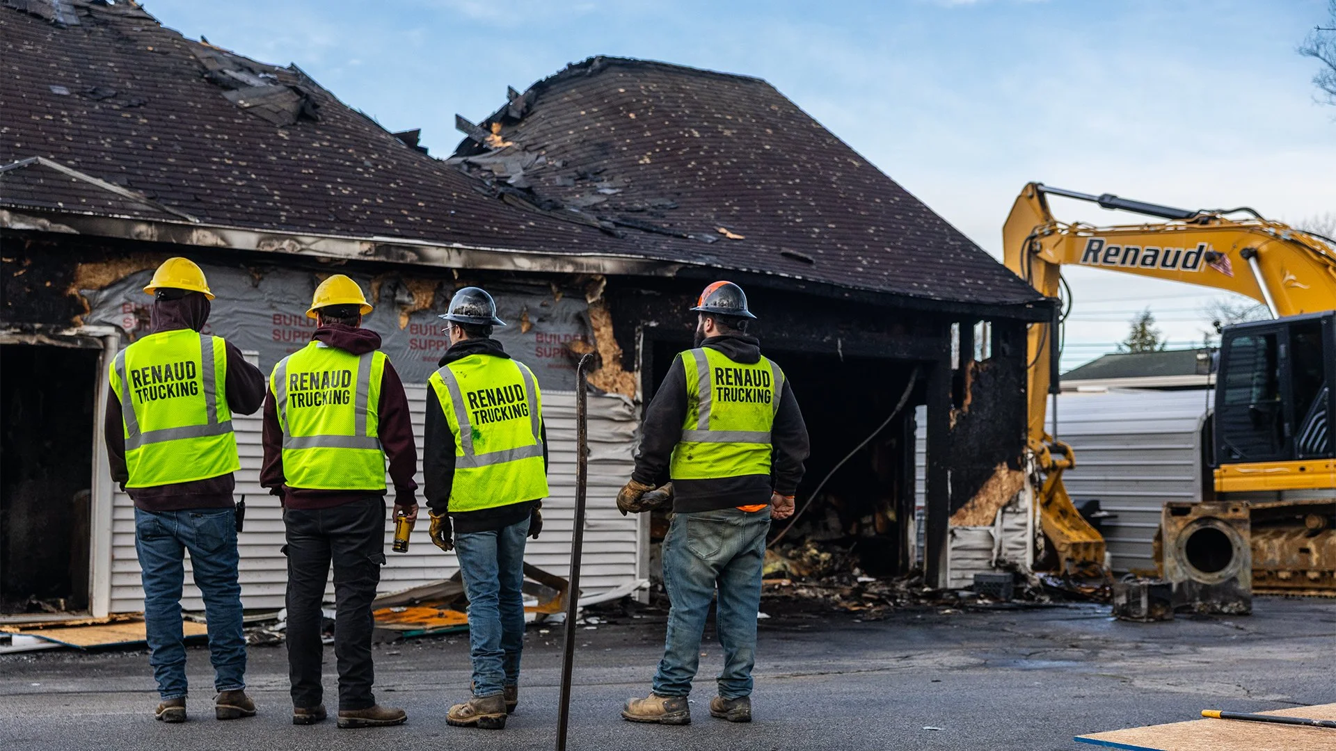 Four construction workers wearing yellow safety vests and hard hats stand in front of a house that has been damaged by fire, with parts of the roof burnt and collapsed, and construction equipment nearby.