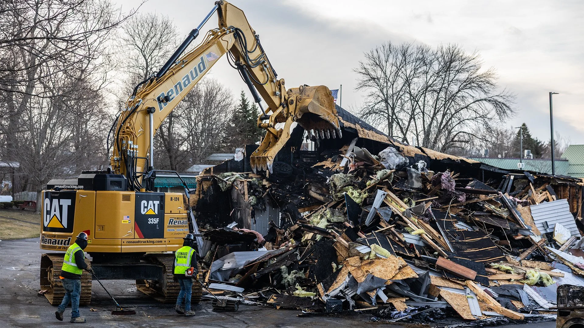 A large CAT construction vehicle with a long arm is demolishing a building, with debris scattered around. Two workers in safety vests are standing nearby observing the demolition.