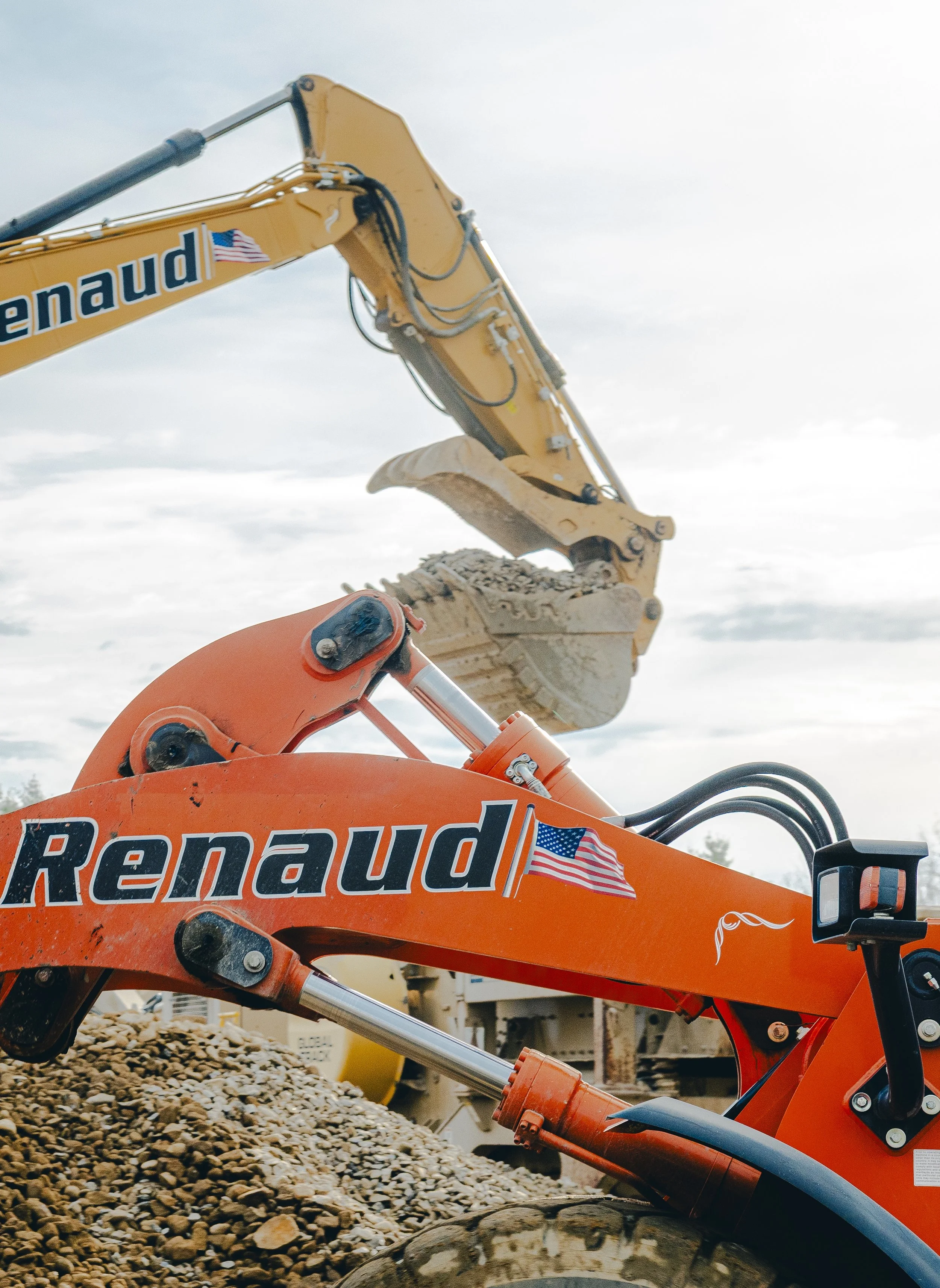 Close-up of an orange Rennaudi-branded construction excavator with a yellow arm and bucket, positioned over a pile of small rocks, under a cloudy sky.
