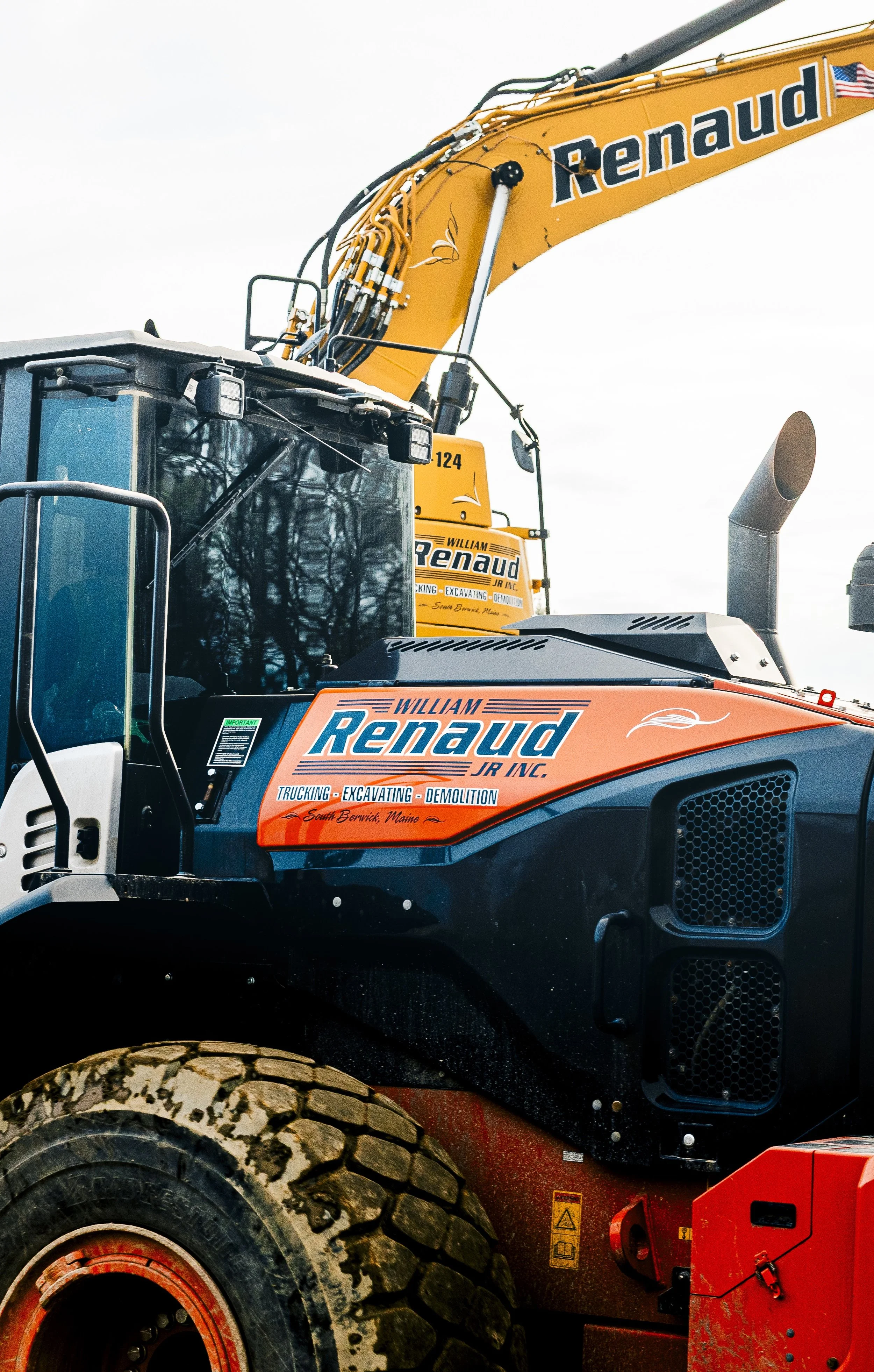 Close-up of a large construction excavator with orange, black, and gray colors, displaying the name 'Renaud Jr. Inc.' along with text indicating truckings, excavating, and demolition services, and a set of large tires with mud on them.