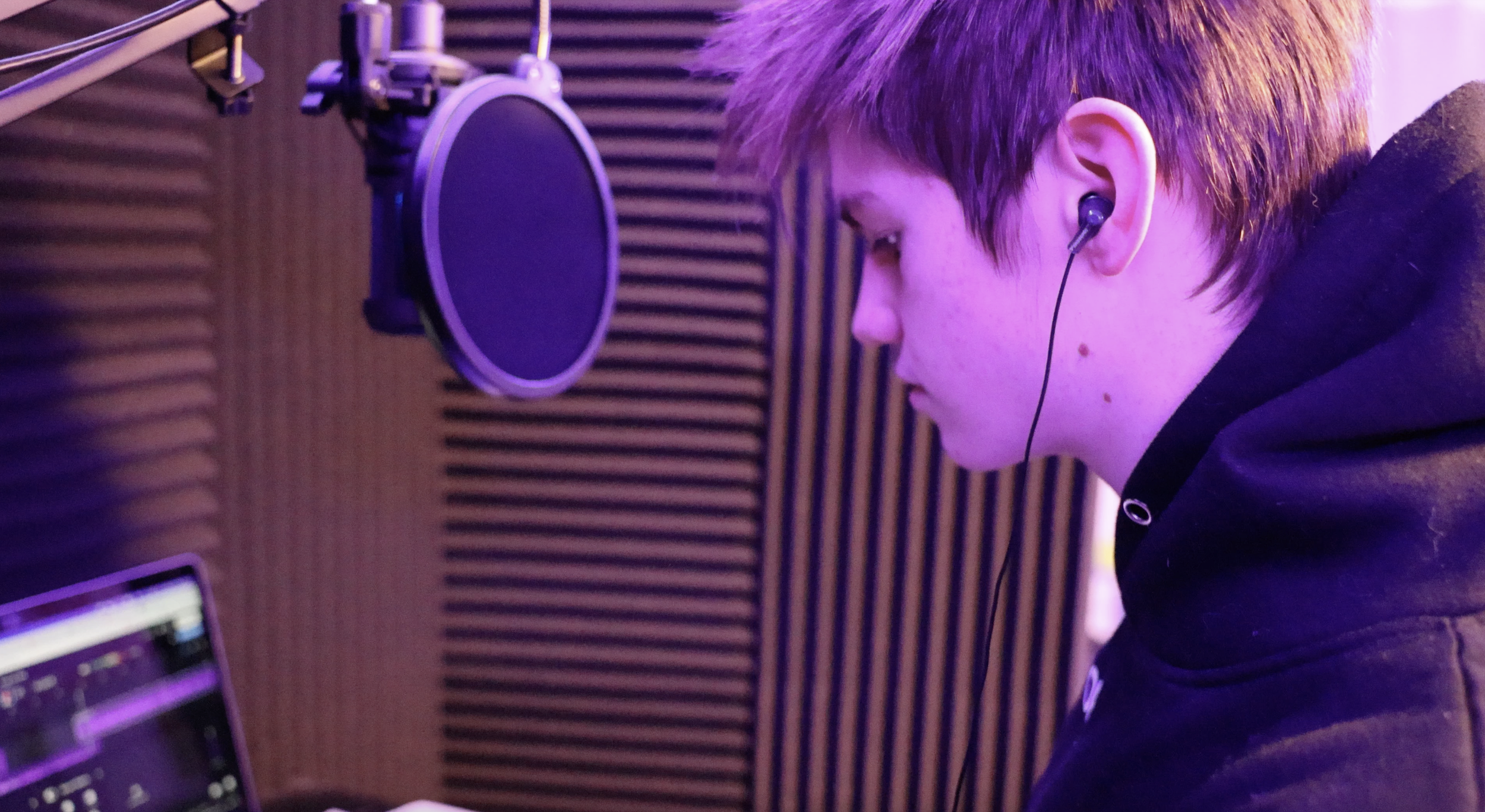 A young person with short hair wearing earphones is in a recording studio facing a computer monitor, with soundproofed walls in the background.