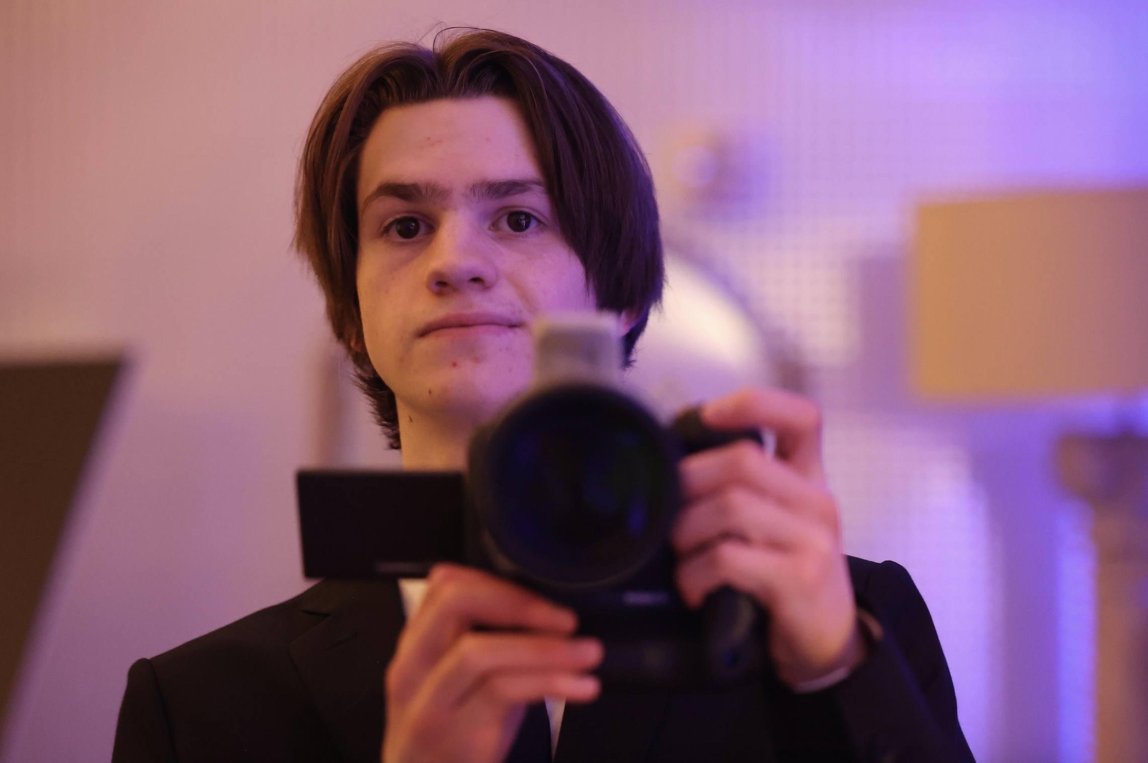 Brennan Martch Media A young man with brown hair taking a selfie with a smartphone in a room illuminated with purple lighting, wearing a dark jacket.