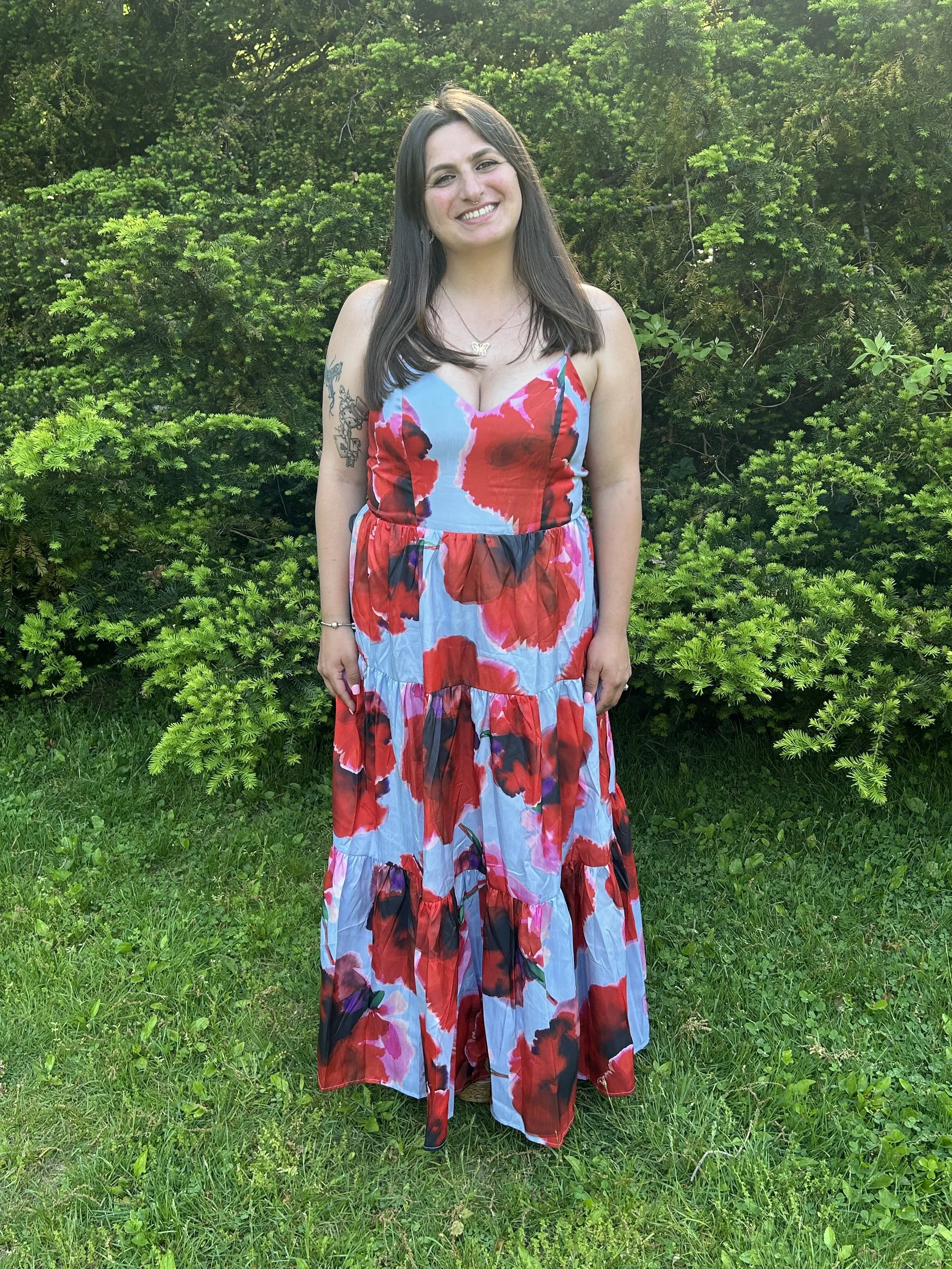 A woman with dark hair, wearing a colorful floral dress, standing outdoors in front of green bushes and grass, smiling at the camera.